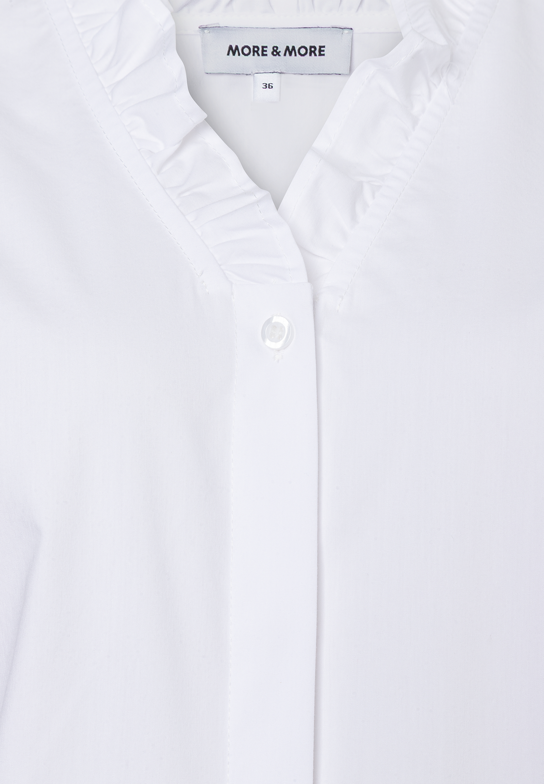 Nahaufnahme einer weißen MORE & MORE Bluse mit Rüschen und Knopf. Nahaufnahme einer weißen MORE & MORE Bluse mit Rüschen und Knopf.