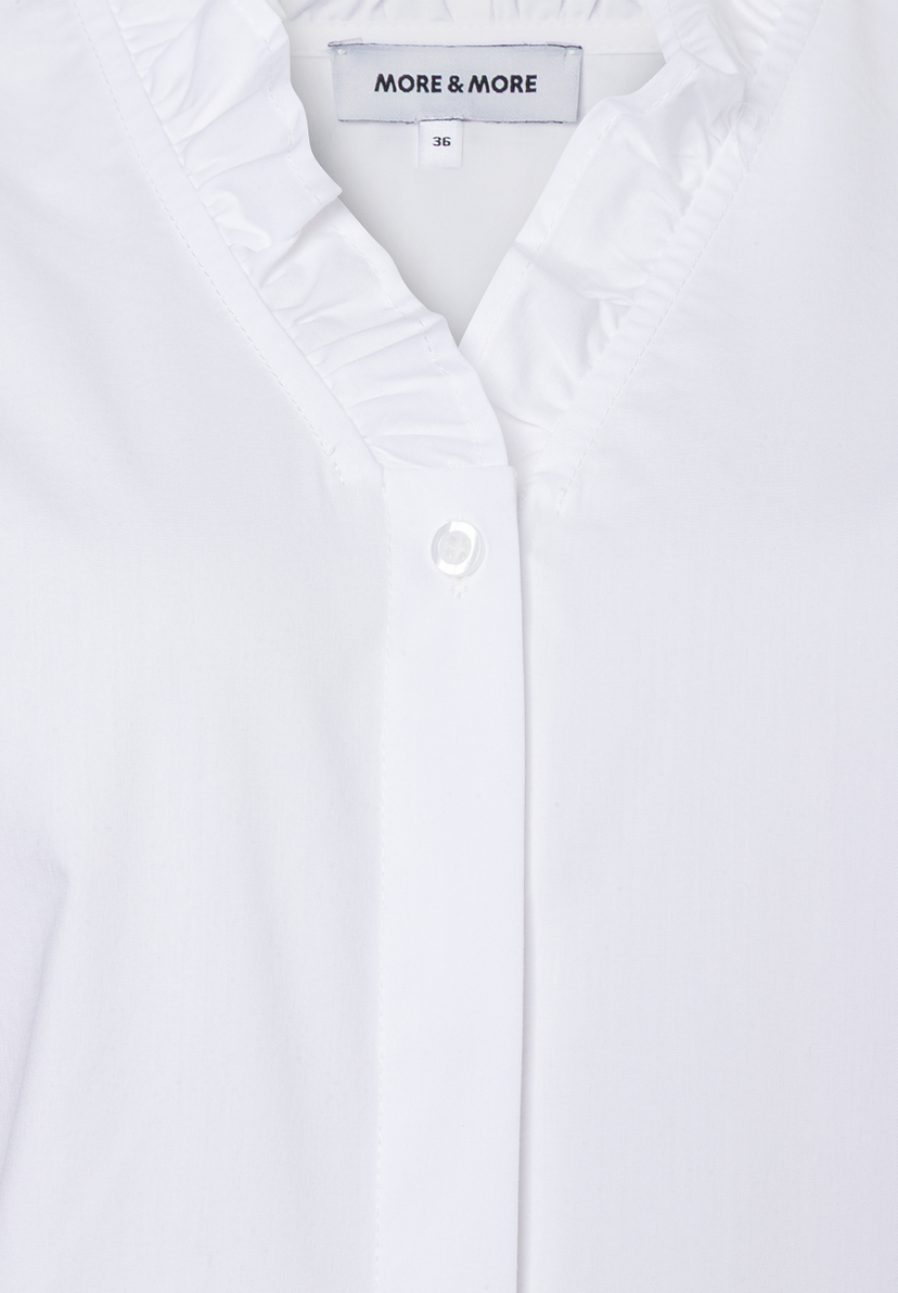 Nahaufnahme einer weißen MORE & MORE Bluse mit Rüschen und Knopf. Nahaufnahme einer weißen MORE & MORE Bluse mit Rüschen und Knopf.
