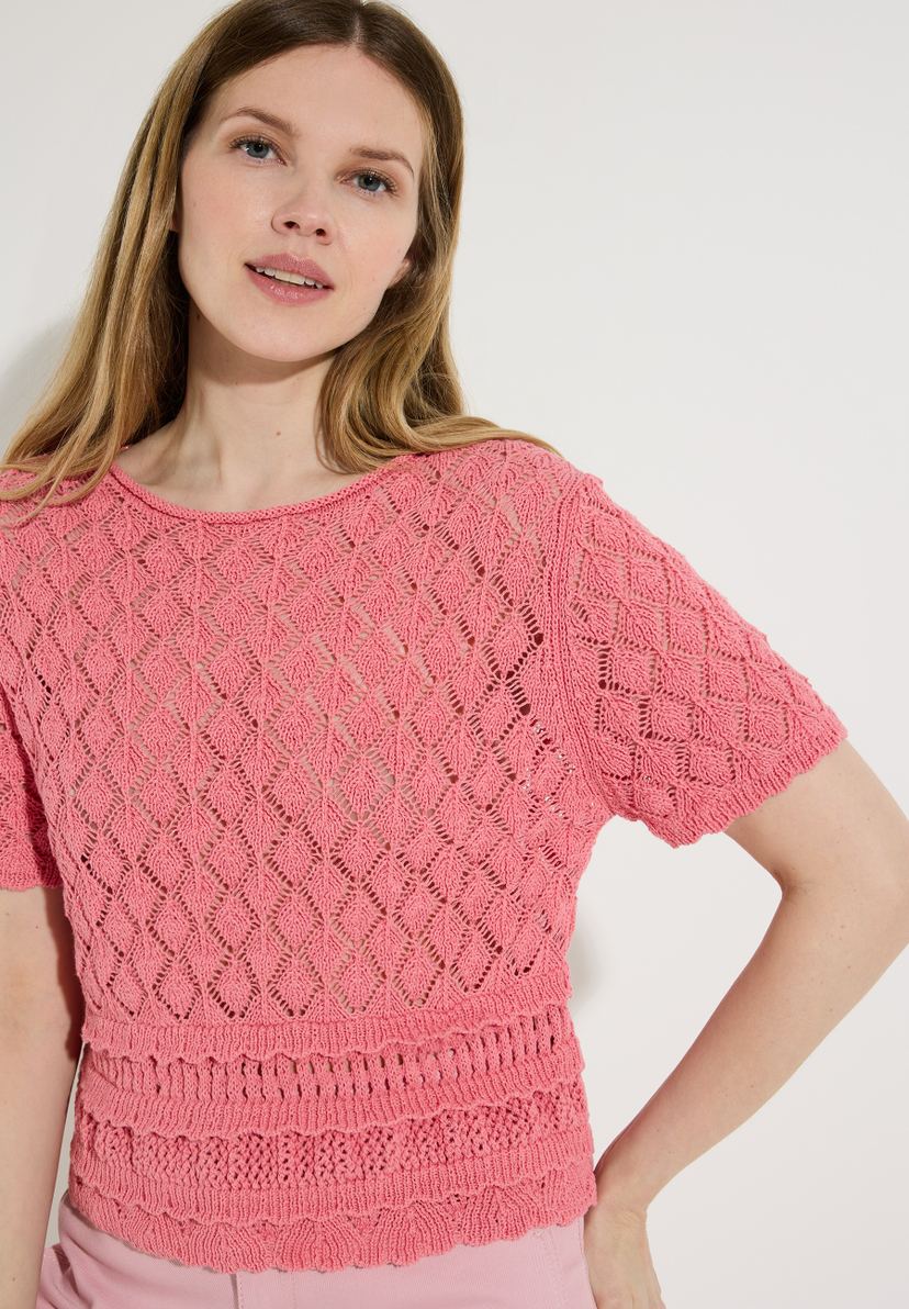 Eine Frau trägt ein korallenrotes Lochstrick-Oberteil der Marke MORE & MORE, das einen leichten Tragekomfort bietet. Eine Frau trägt ein korallenrotes Lochstrick-Oberteil der Marke MORE & MORE, das einen leichten Tragekomfort bietet.