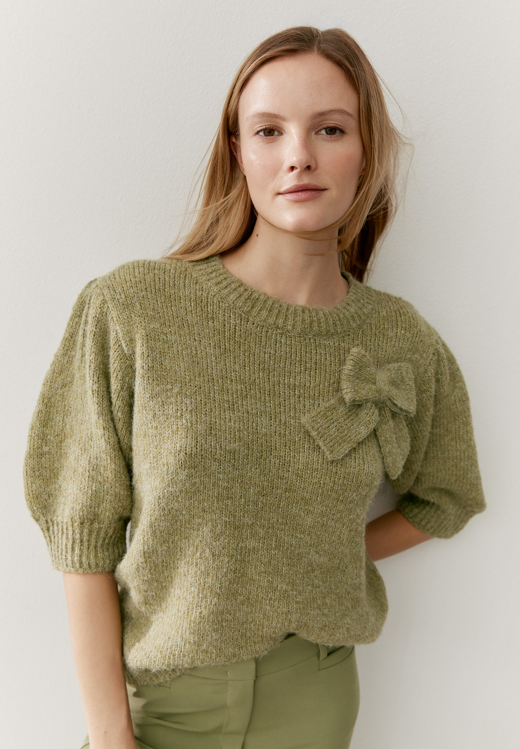 Eine Frau trägt einen grünen Strickpullover mit Schleife von MORE & MORE.