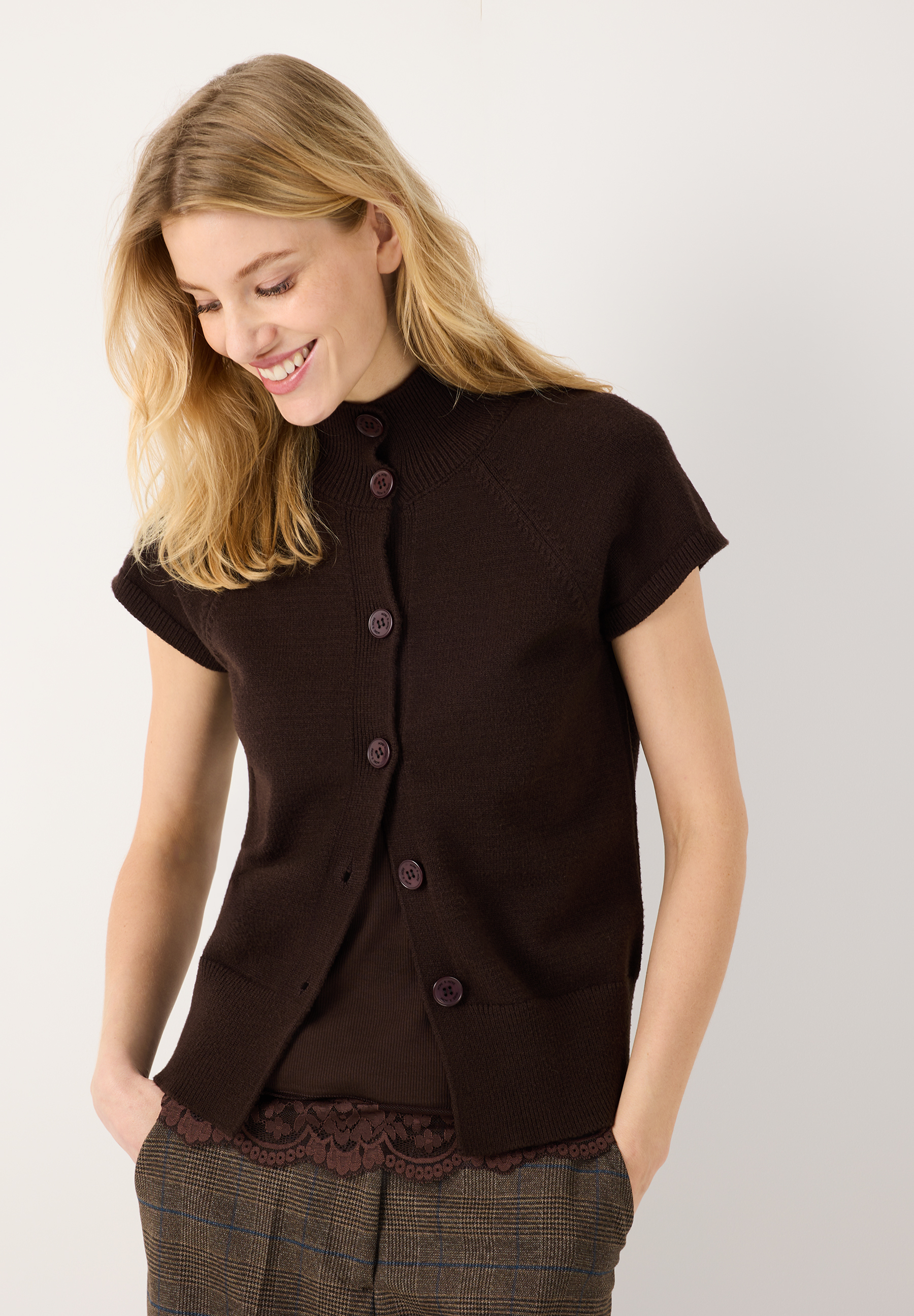MORE & MORE Pullover: Eine lächelnde blonde Frau in einem schokobraunen Strick-Kurzarmpullover, die Hände in den Hosentaschen. 