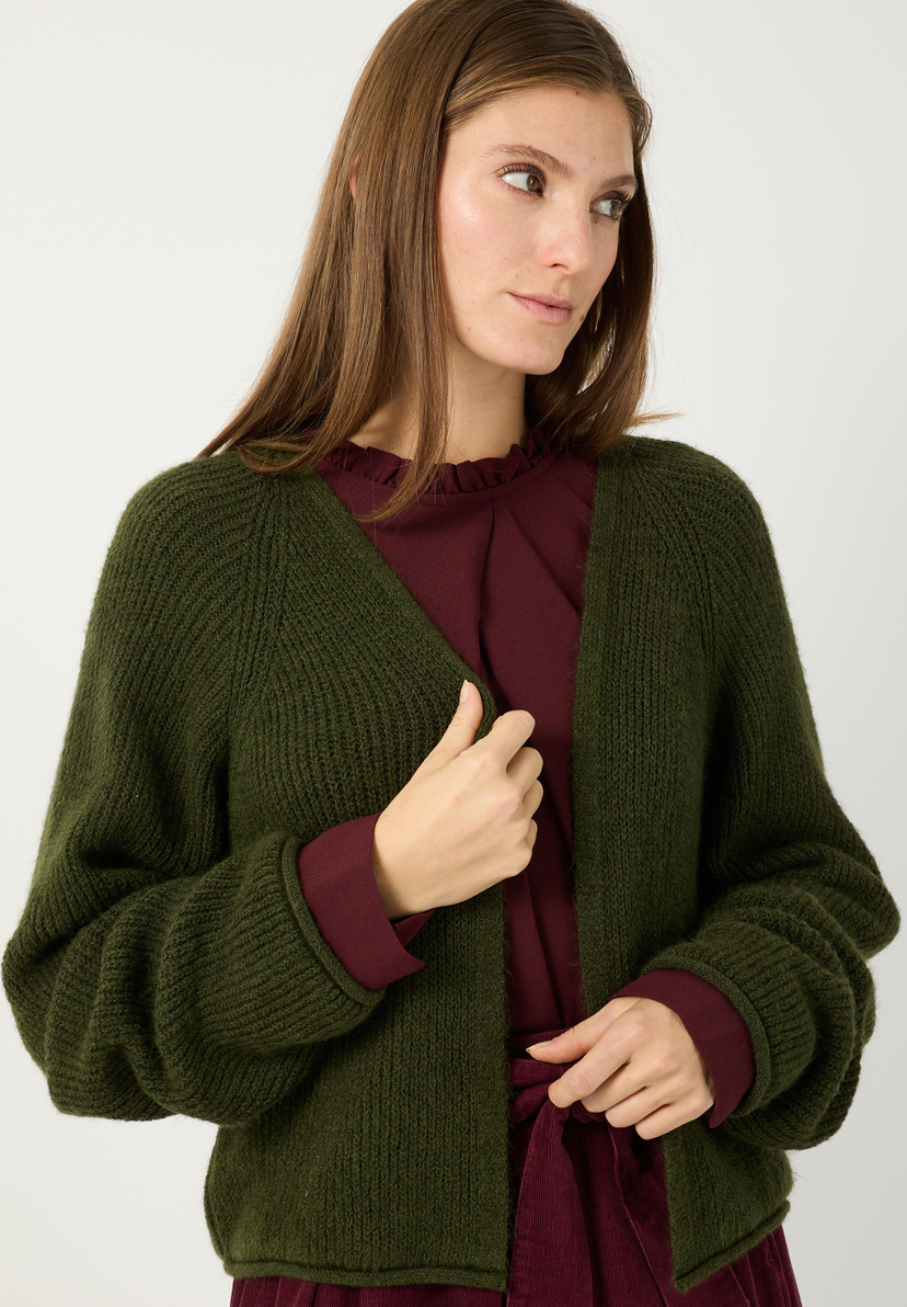 MORE & MORE Strickjacke: Eine Frau trägt eine olivgrüne Strickjacke und ein weinrotes Oberteil. Ihre Hände sind vor ihr verschränkt.