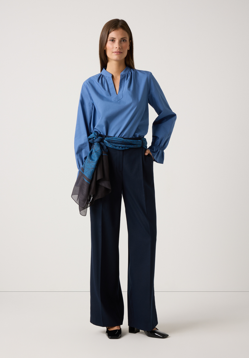 Eine Frau trägt eine blaue MORE & MORE Bluse, eine dunkle Hose und einen Schal als Gürtel vor einem hellen Hintergrund. Eine Frau trägt eine blaue MORE & MORE Bluse, eine dunkle Hose und einen Schal als Gürtel vor einem hellen Hintergrund.