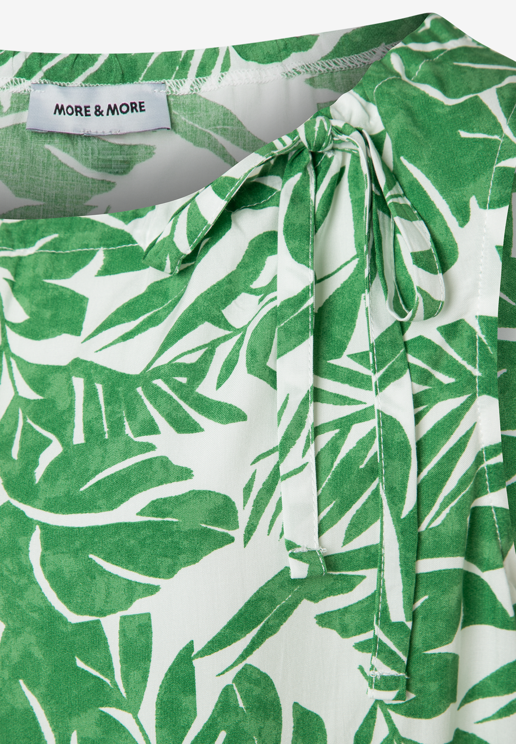 Nahaufnahme einer MORE & MORE Bluse mit grünem Blätterdruck und Schleifendetail am Ausschnitt. Nahaufnahme einer MORE & MORE Bluse mit grünem Blätterdruck und Schleifendetail am Ausschnitt.