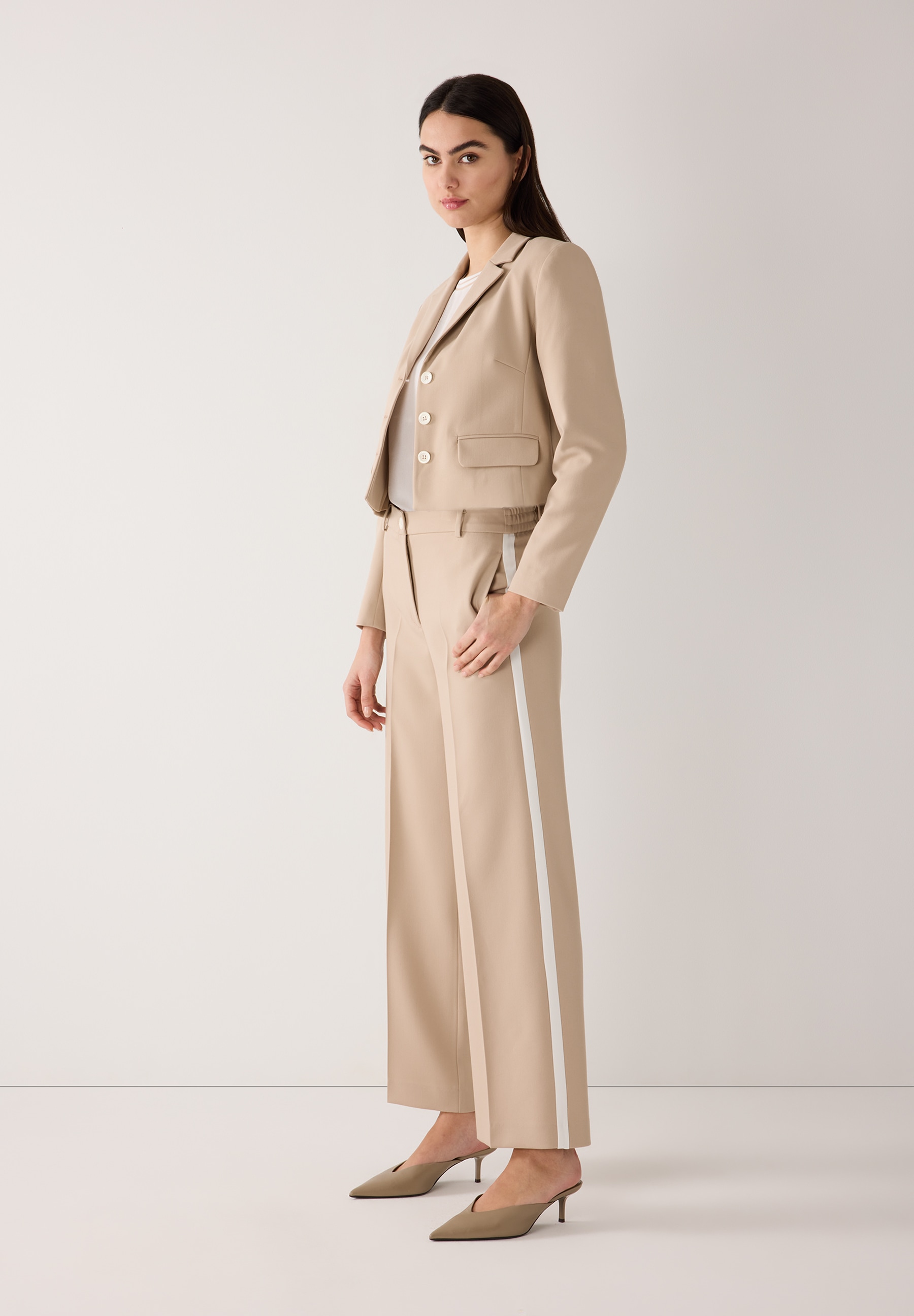 Eine Frau im MORE & MORE Blazer-Ensemble mit beigen Hosen mit weißen Streifen und passenden Mules. Eine Frau im MORE & MORE Blazer-Ensemble mit beigen Hosen mit weißen Streifen und passenden Mules.