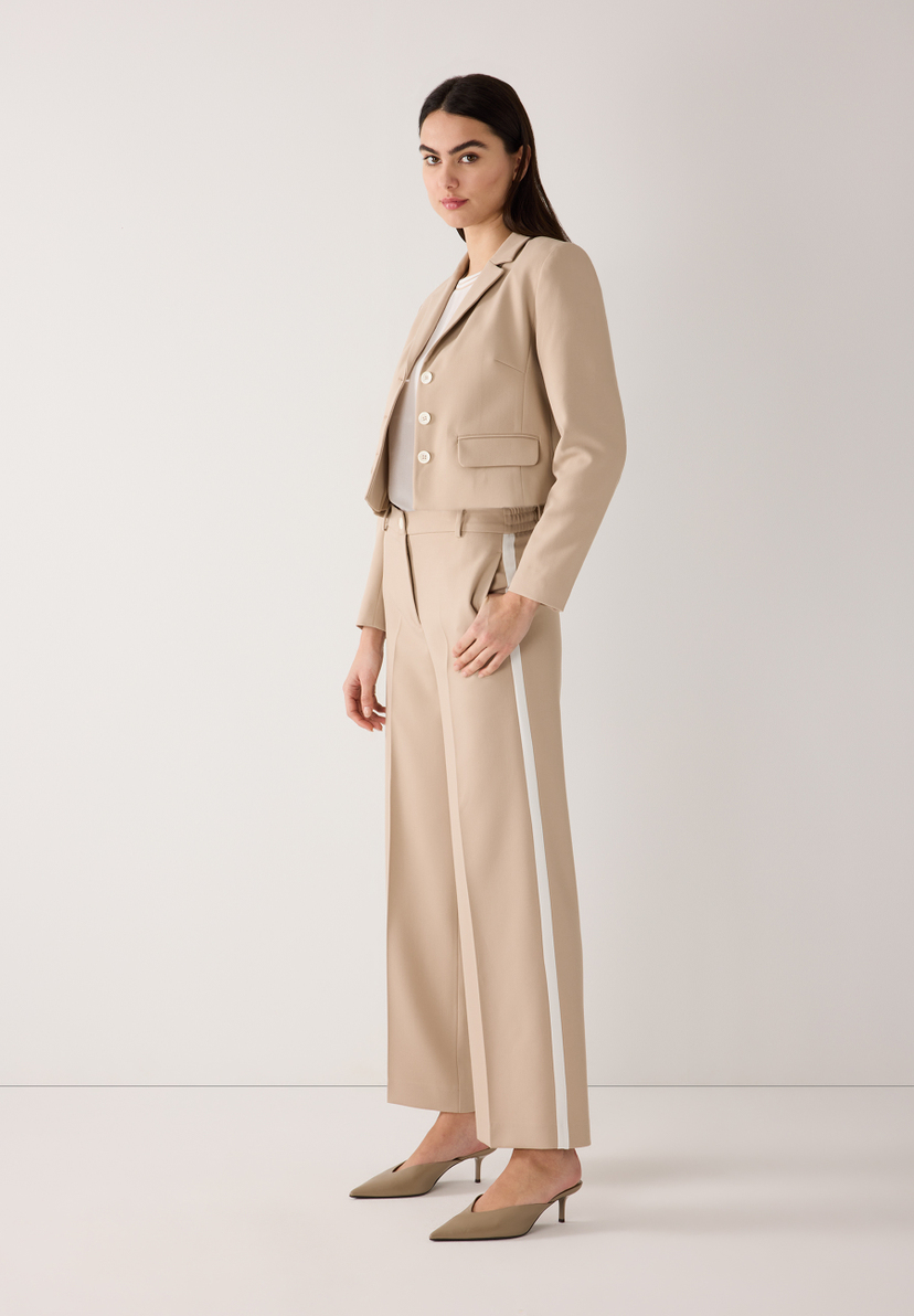 Eine Frau im MORE & MORE Blazer-Ensemble mit beigen Hosen mit weißen Streifen und passenden Mules. Eine Frau im MORE & MORE Blazer-Ensemble mit beigen Hosen mit weißen Streifen und passenden Mules.