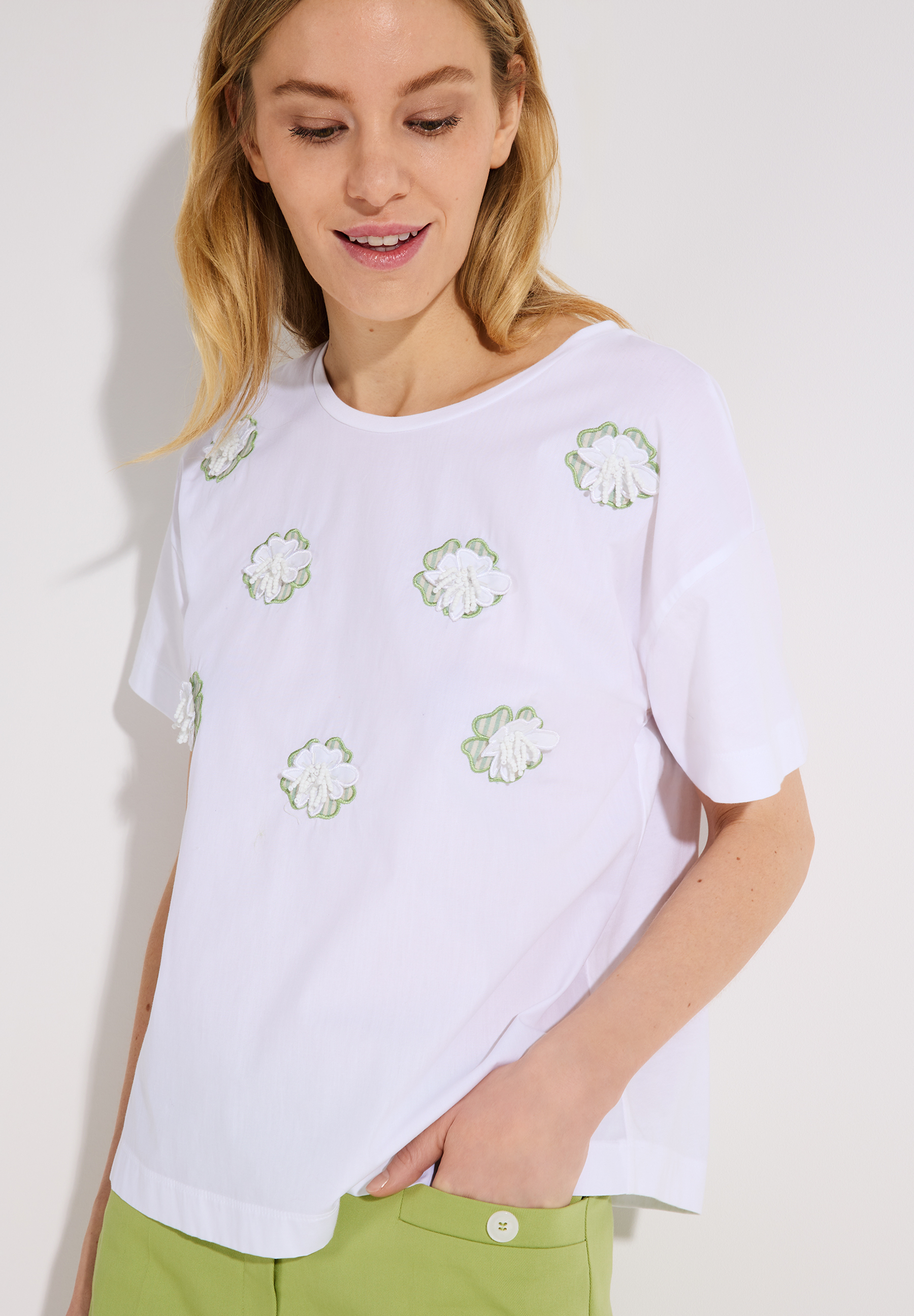 Eine Frau mit einem weißen MORE & MORE T-Shirt mit Blumenapplikationen. Eine Frau mit einem weißen MORE & MORE T-Shirt mit Blumenapplikationen.