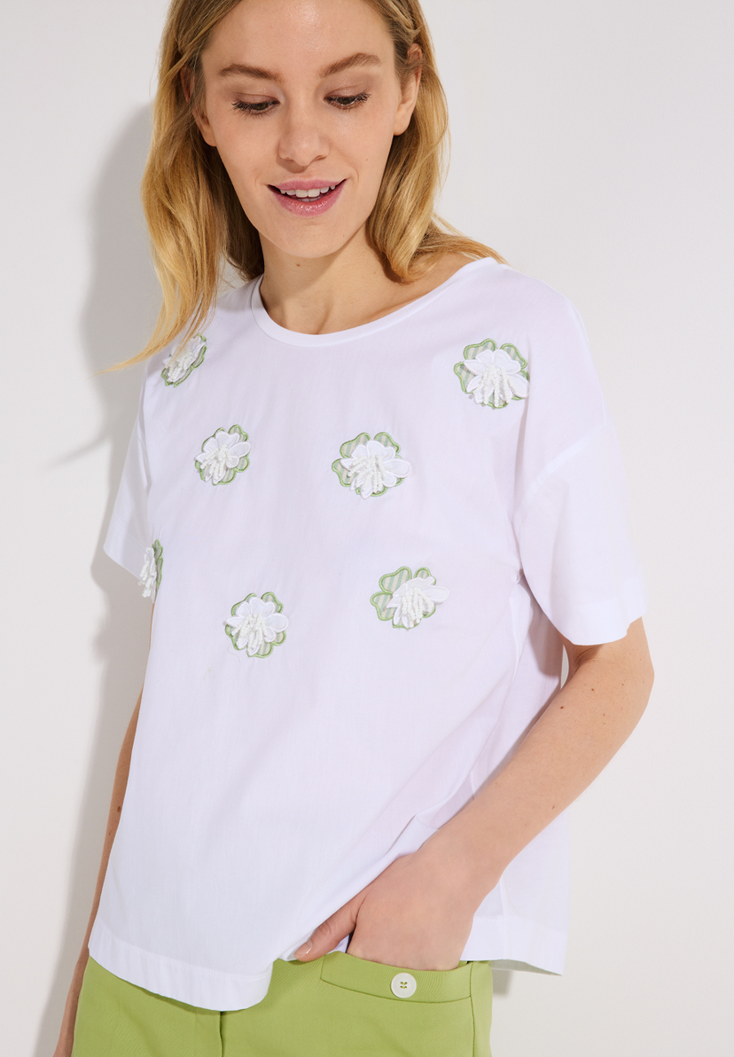 Eine Frau mit einem weißen MORE & MORE T-Shirt mit Blumenapplikationen. Eine Frau mit einem weißen MORE & MORE T-Shirt mit Blumenapplikationen.