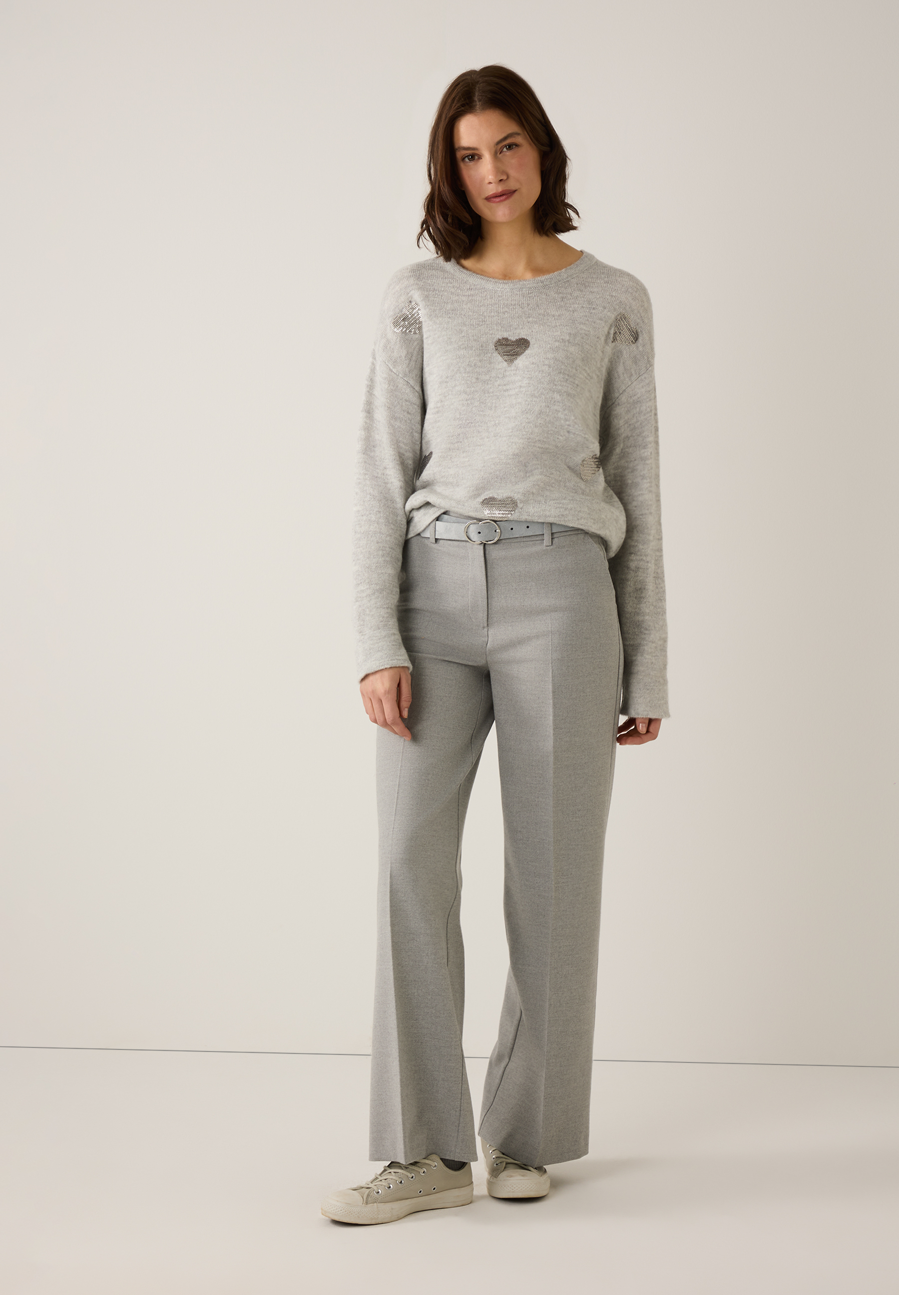 Eine Frau trägt einen grauen MORE & MORE Feinstrick-Pullover mit Herzapplikationen und eine graue Stoffhose mit passendem Gürtel. Eine Frau trägt einen grauen MORE & MORE Feinstrick-Pullover mit Herzapplikationen und eine graue Stoffhose mit passendem Gürtel.