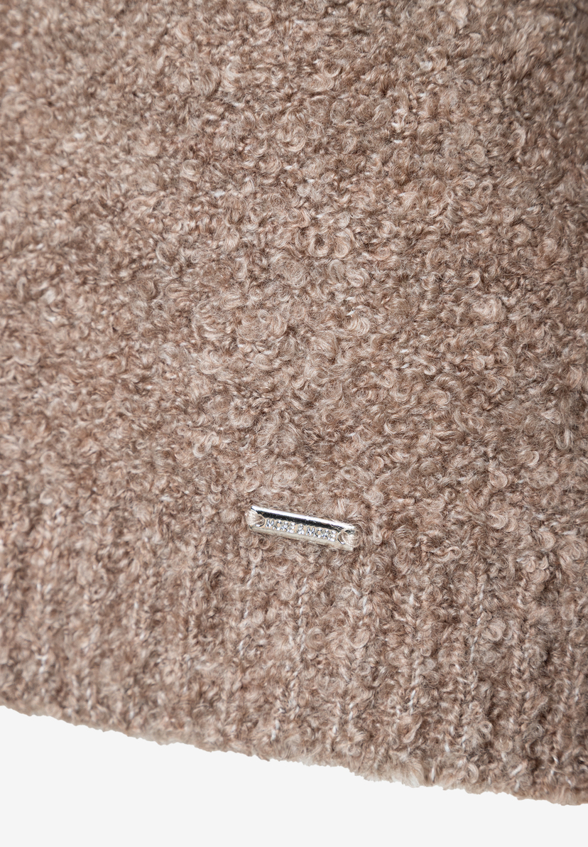 Nahaufnahme eines warmen, beigen Bouclé-Pullovers von MORE & MORE mit Logo-Metallplakette. Nahaufnahme eines warmen, beigen Bouclé-Pullovers von MORE & MORE mit Logo-Metallplakette.