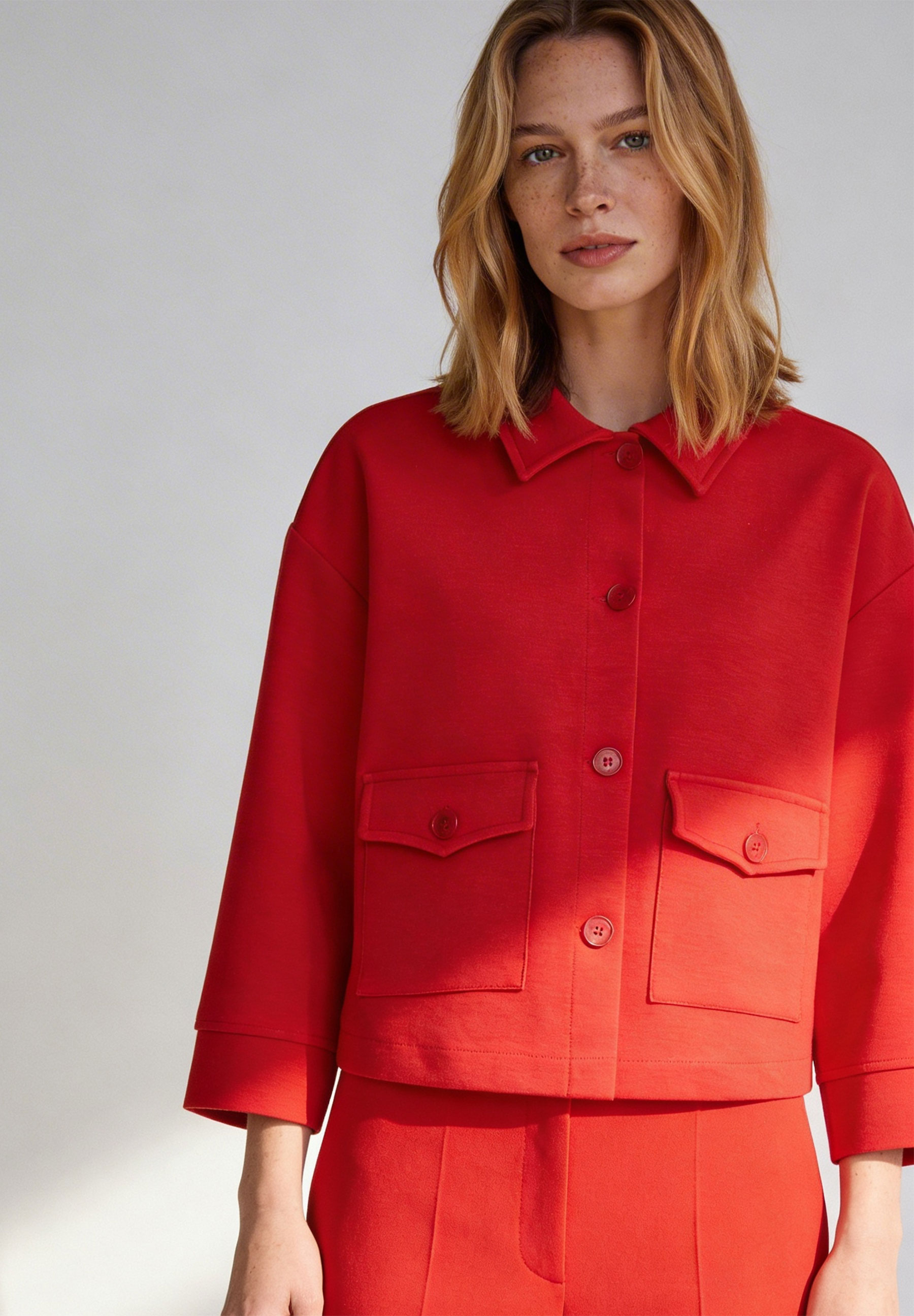 Eine Frau mit einem roten MORE & MORE Jersey-Oberteil mit Knopfleiste und zwei aufgesetzten Taschen.