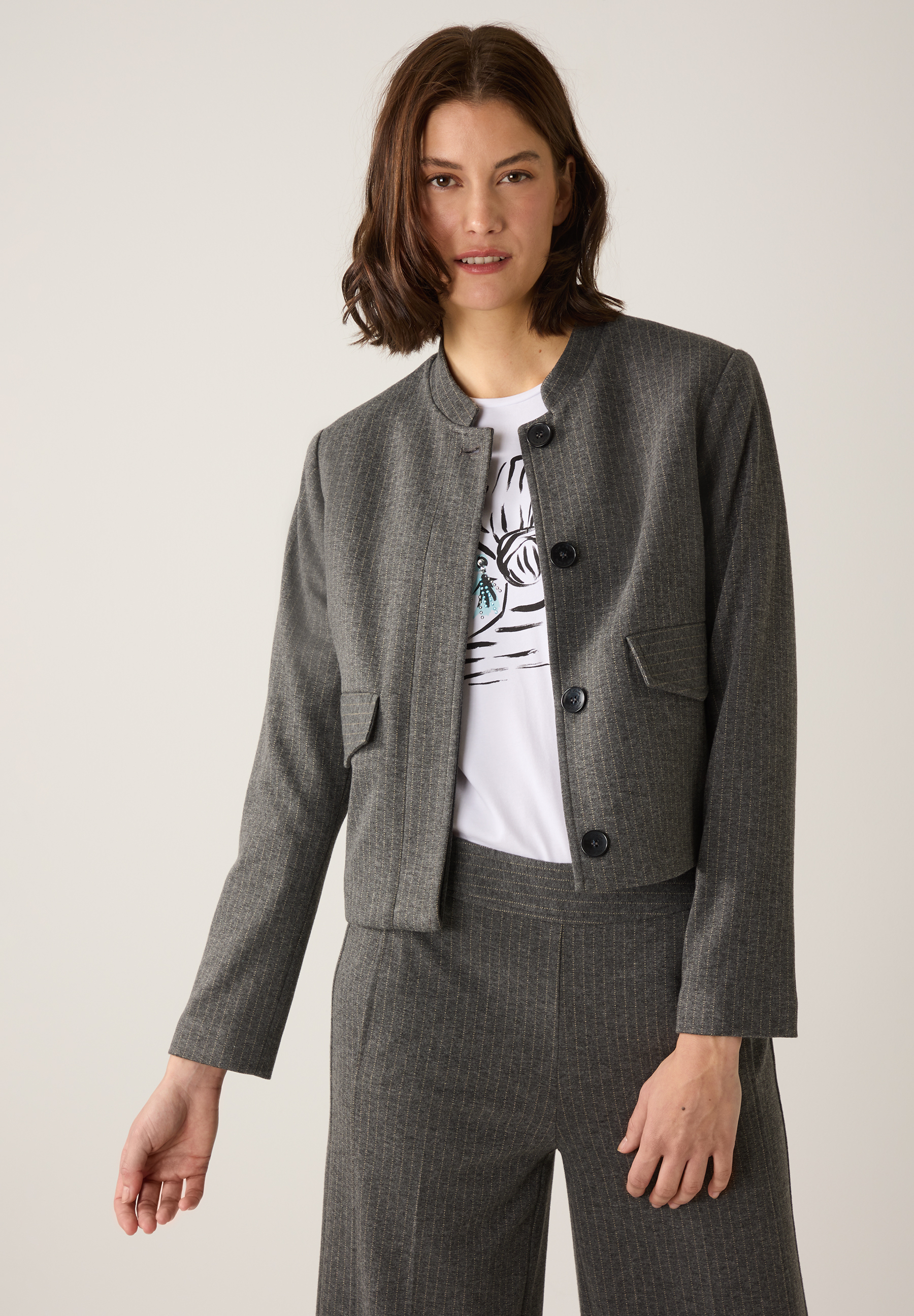 Eine Frau trägt einen grauen MORE & MORE Blazer mit Nadelstreifen über einem weißen T-Shirt. Eine Frau trägt einen grauen MORE & MORE Blazer mit Nadelstreifen über einem weißen T-Shirt.