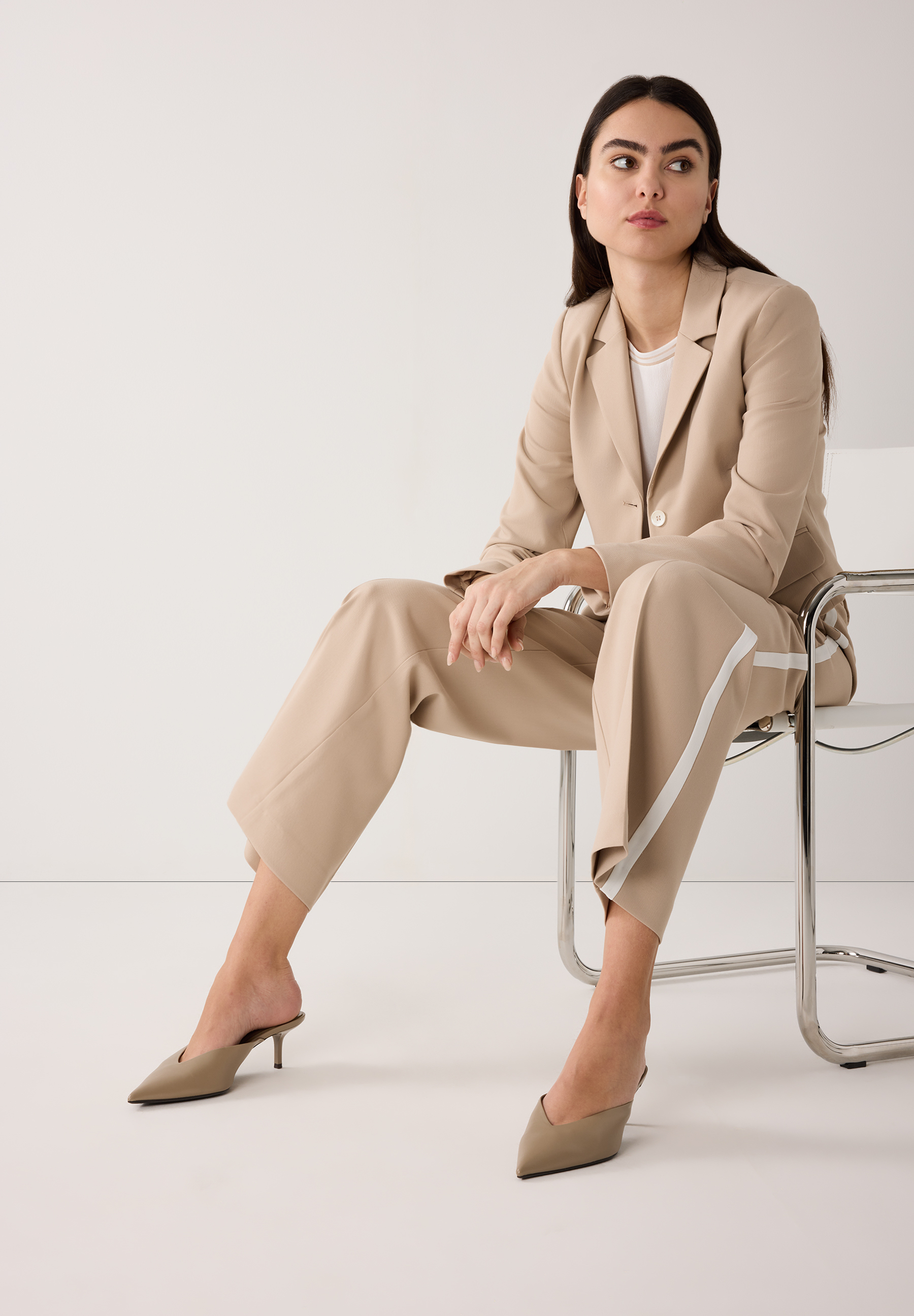 Eine Frau sitzt auf einem Stuhl und trägt einen MORE & MORE Blazer und passende Hose. Die Kleidung ist hellbeige. Eine Frau sitzt auf einem Stuhl und trägt einen MORE & MORE Blazer und passende Hose. Die Kleidung ist hellbeige.