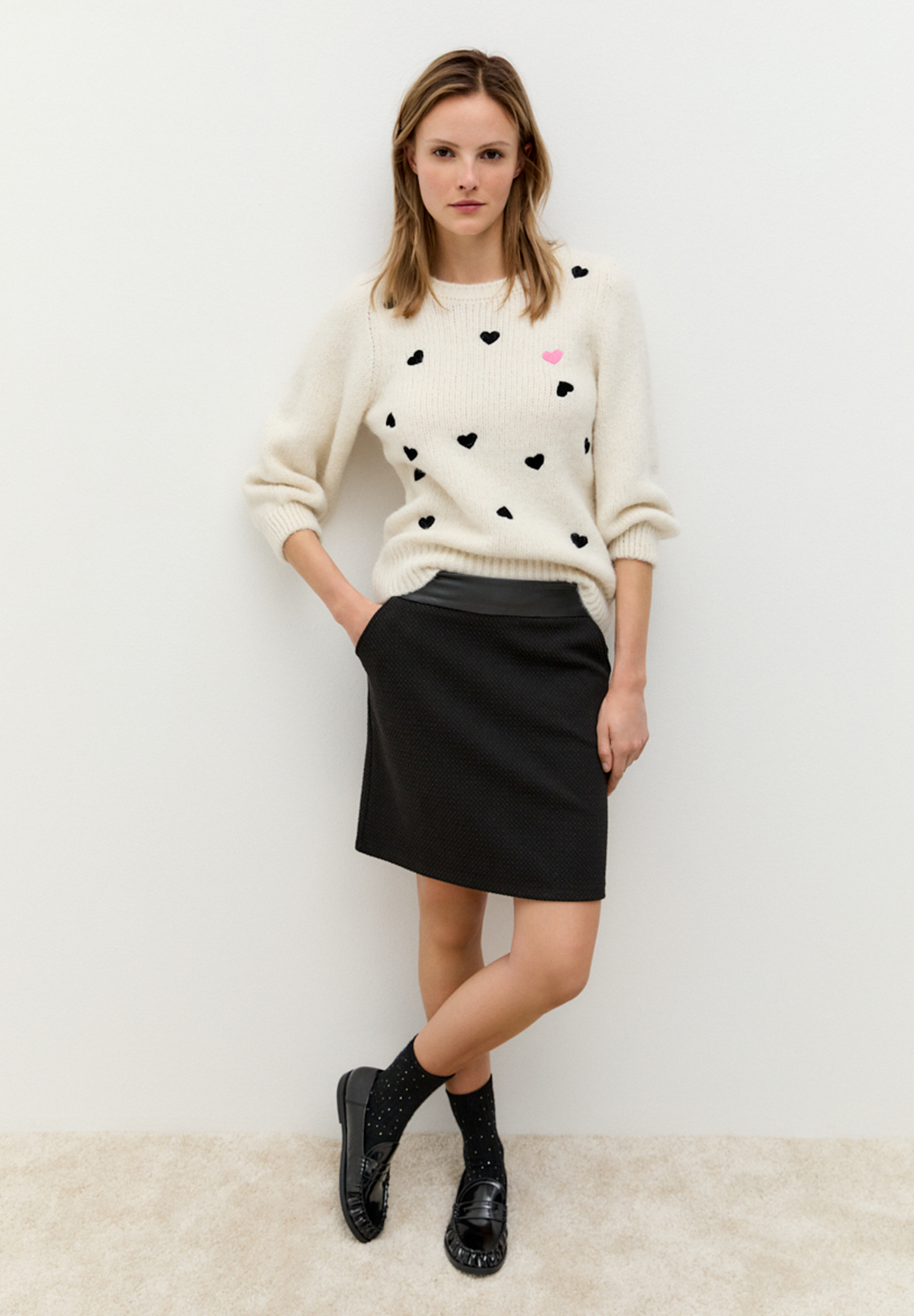 Eine Frau posiert in einem MORE & MORE Oberteil, einem cremefarbenen Pullover mit schwarzen und einem rosa Herzmuster.