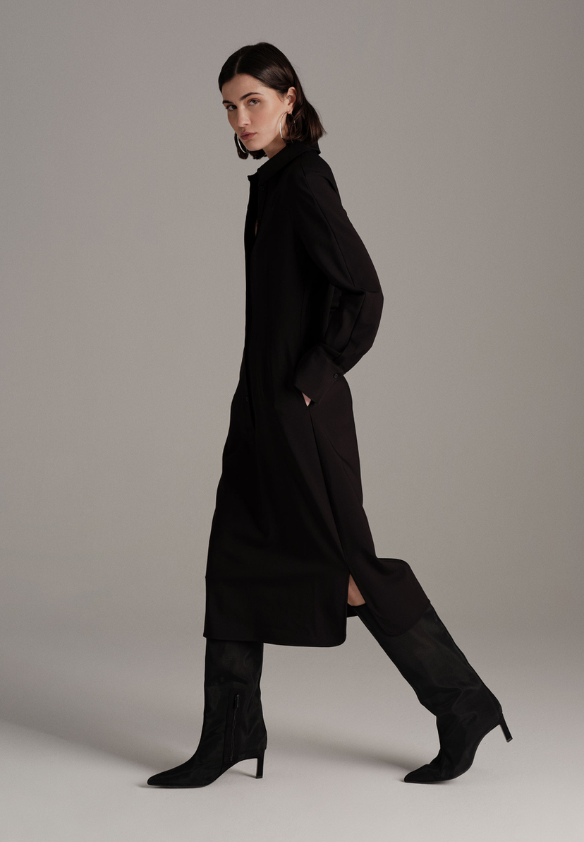 Eine Frau in einem schwarzen MORE & MORE Oberteil-Kleid und Stiefeln posiert seitlich vor grauem Hintergrund. Eine Frau in einem schwarzen MORE & MORE Oberteil-Kleid und Stiefeln posiert seitlich vor grauem Hintergrund.