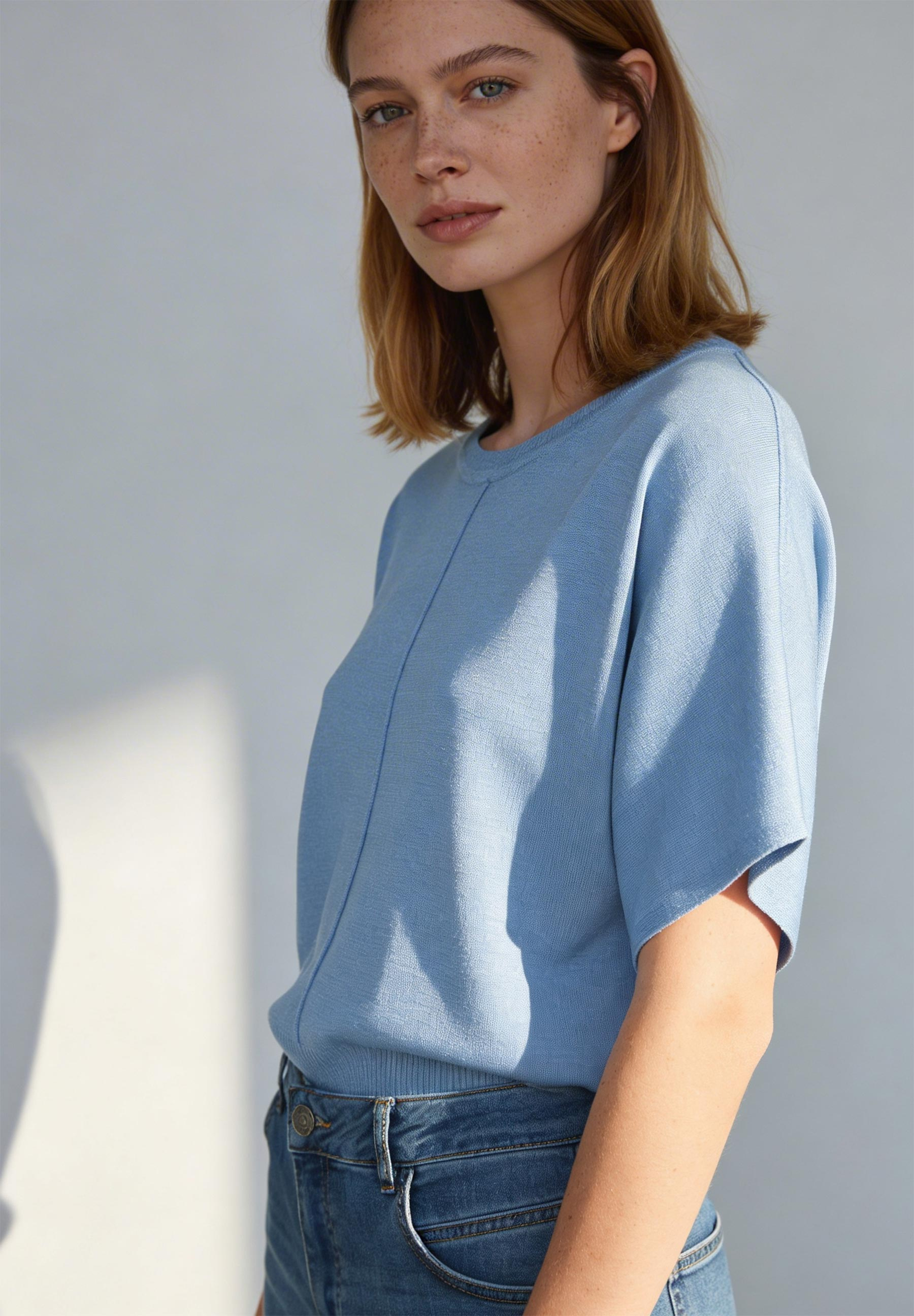 Eine Frau trägt ein hellblaues Oberteil der Marke MORE & MORE mit Jeans vor einer hellen Wand.