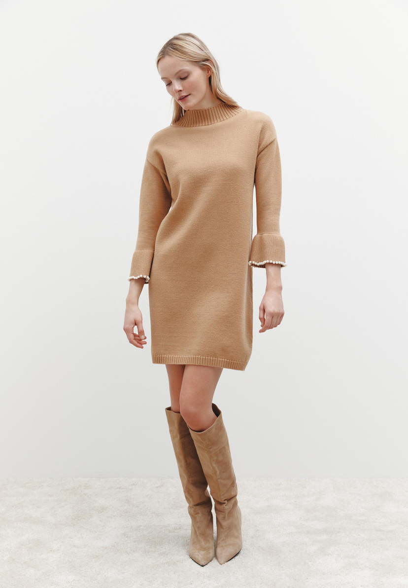 Eine blonde Frau in einem beigen MORE & MORE Strickkleid und Stiefeln vor einem weißen Hintergrund. Eine blonde Frau in einem beigen MORE & MORE Strickkleid und Stiefeln vor einem weißen Hintergrund.