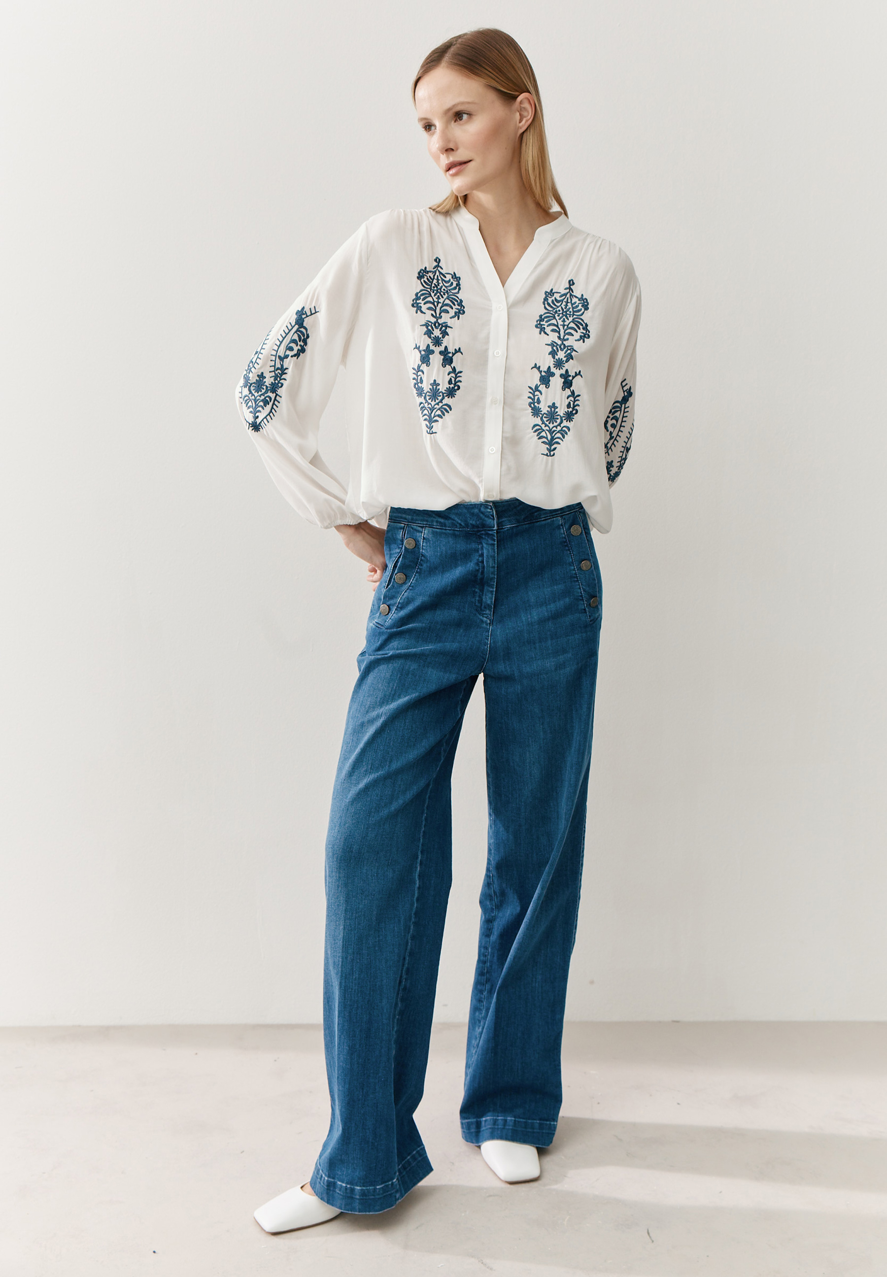 Eine Frau trägt eine weiße MORE & MORE Bluse mit blauen Stickereien und Jeans. Eine Frau trägt eine weiße MORE & MORE Bluse mit blauen Stickereien und Jeans.