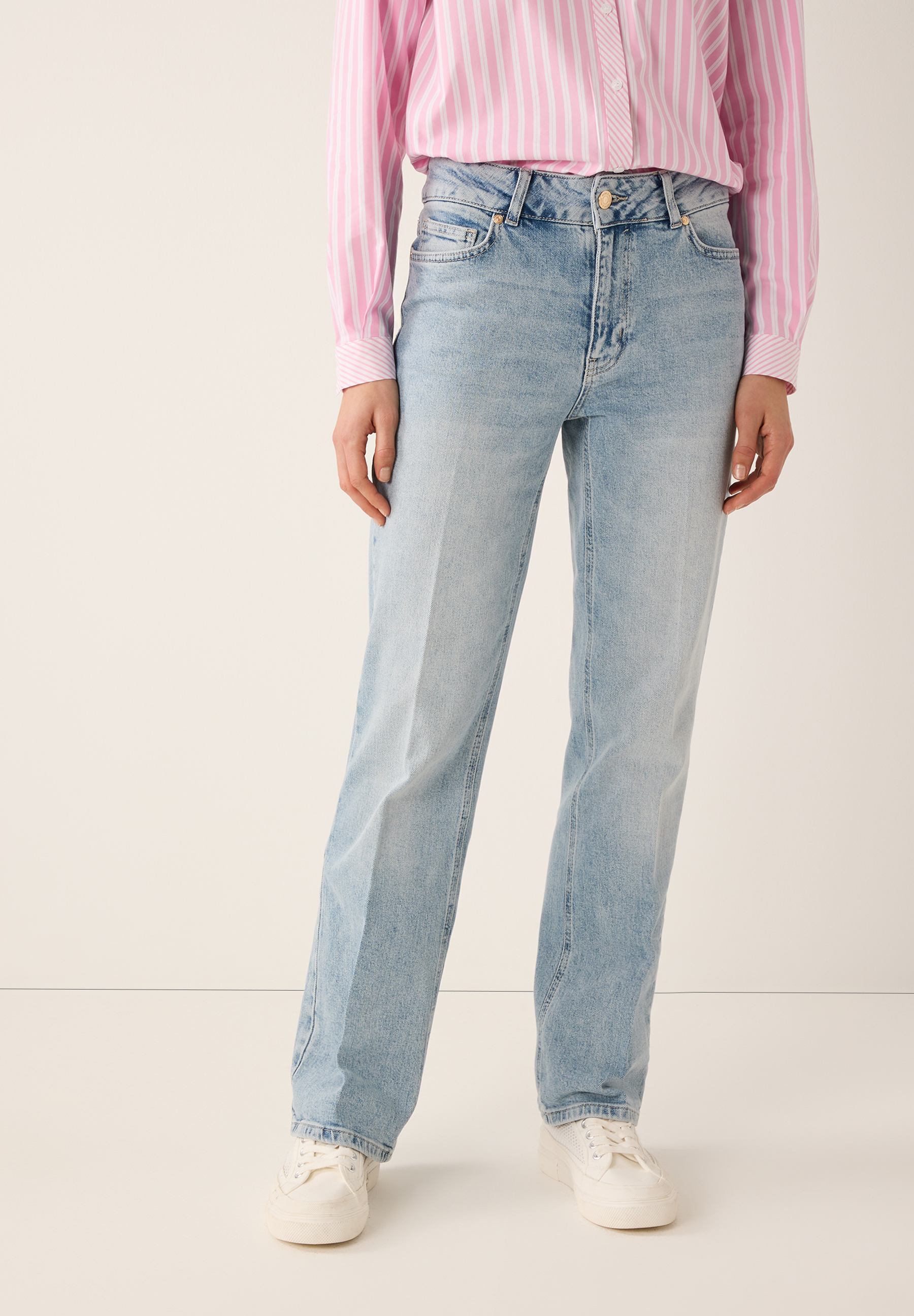 Eine Person trägt MORE & MORE Jeans mit rosa-weiß gestreiftem Hemd und weißen Turnschuhen vor einem schlichten Hintergrund.