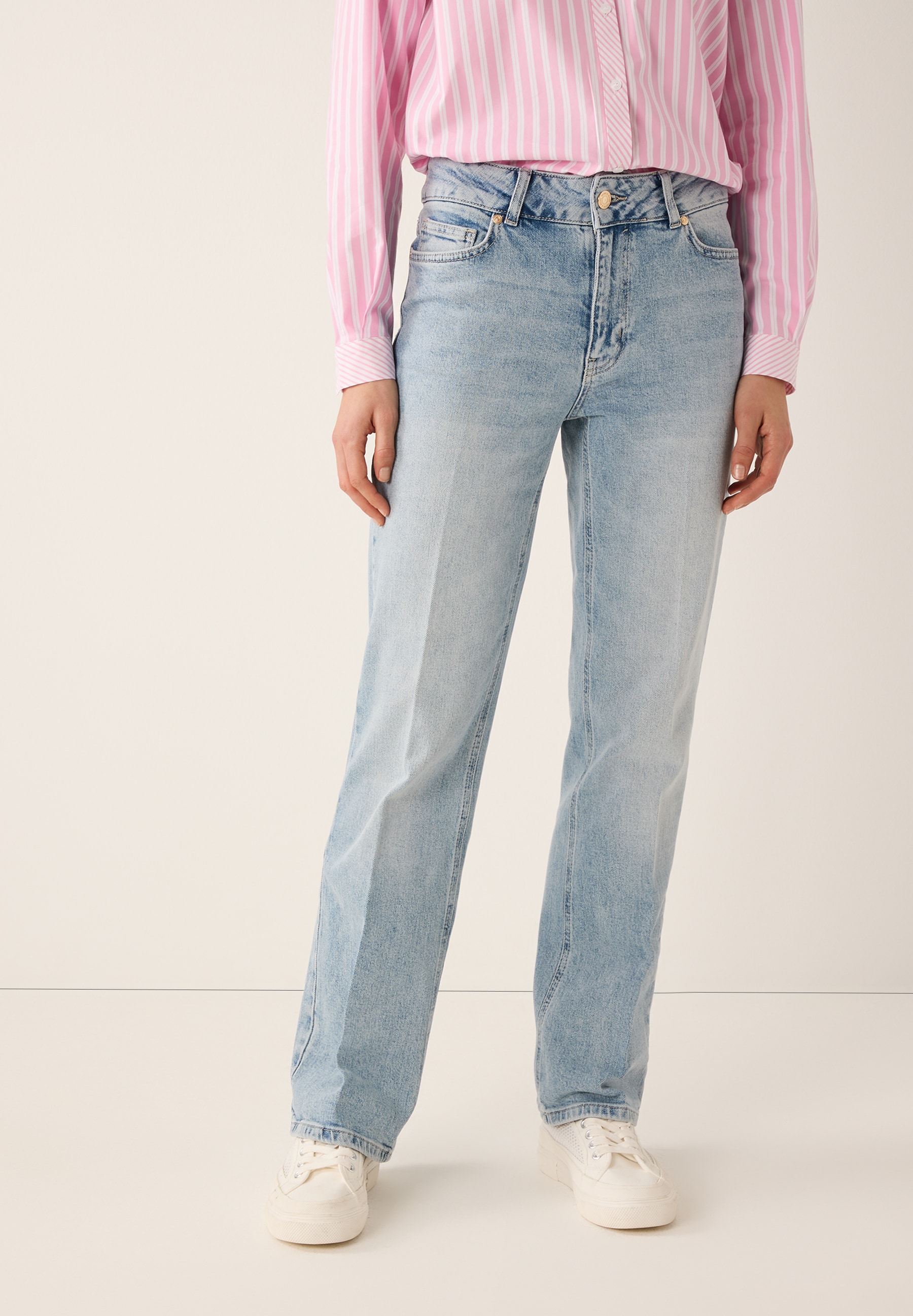 Eine Person trägt MORE & MORE Jeans mit rosa-weiß gestreiftem Hemd und weißen Turnschuhen vor einem schlichten Hintergrund.