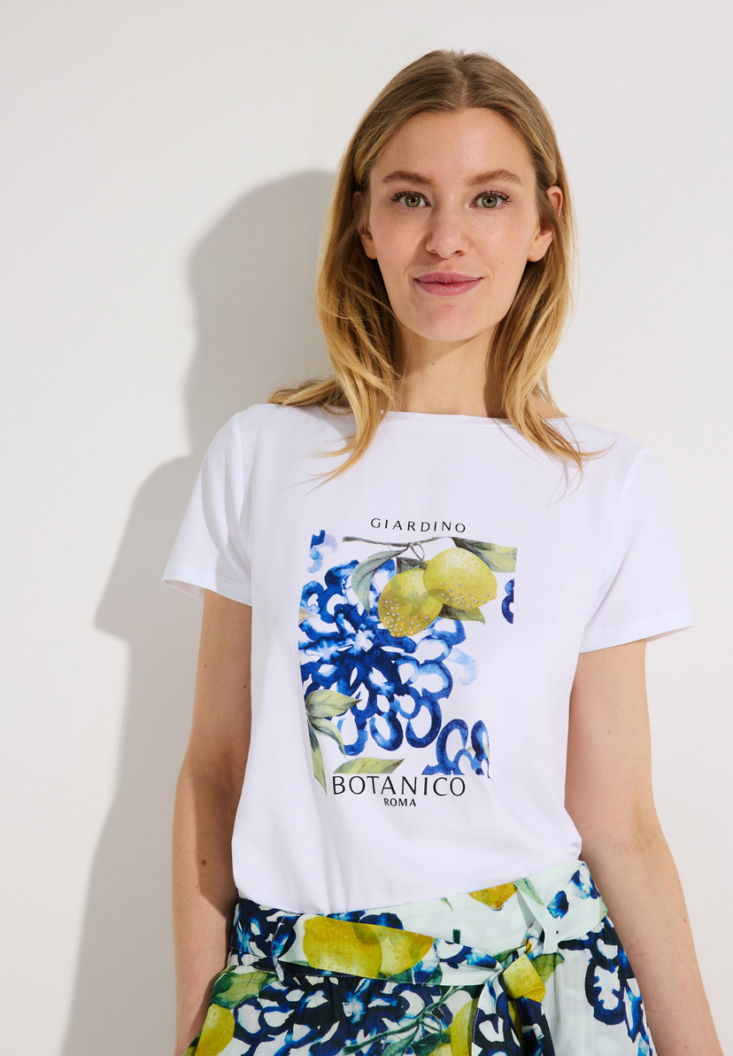 Eine Frau trägt ein weißes MORE & MORE T-Shirt mit einem Blumen- und Zitronenmotiv. Eine Frau trägt ein weißes MORE & MORE T-Shirt mit einem Blumen- und Zitronenmotiv.