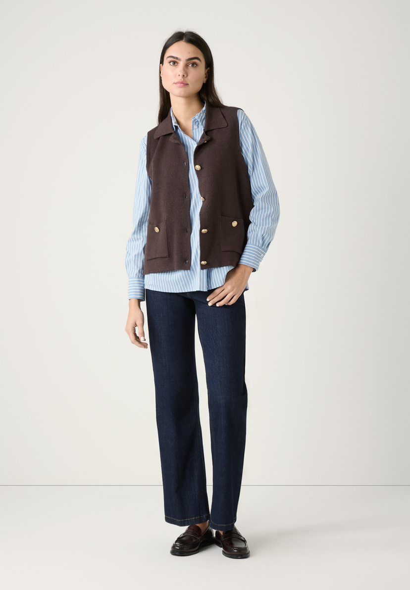 Eine Frau, die MORE & MORE Oberteile, eine Weste über einem Hemd, zu Jeans und Schuhen trägt. Eine Frau, die MORE & MORE Oberteile, eine Weste über einem Hemd, zu Jeans und Schuhen trägt.