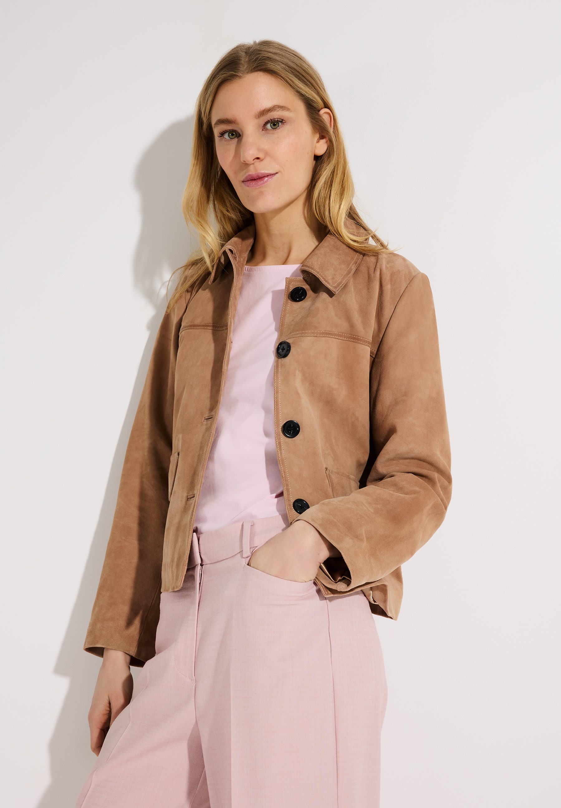 Eine blonde Frau trägt eine MORE & MORE Velourslederjacke über einem rosa Oberteil und einer passenden Hose. Eine blonde Frau trägt eine MORE & MORE Velourslederjacke über einem rosa Oberteil und einer passenden Hose.