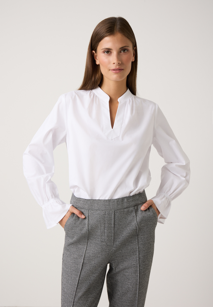Eine Frau mit einer weißen MORE & MORE Bluse und grauen Hosen posiert vor einem weißen Hintergrund. Eine Frau mit einer weißen MORE & MORE Bluse und grauen Hosen posiert vor einem weißen Hintergrund.