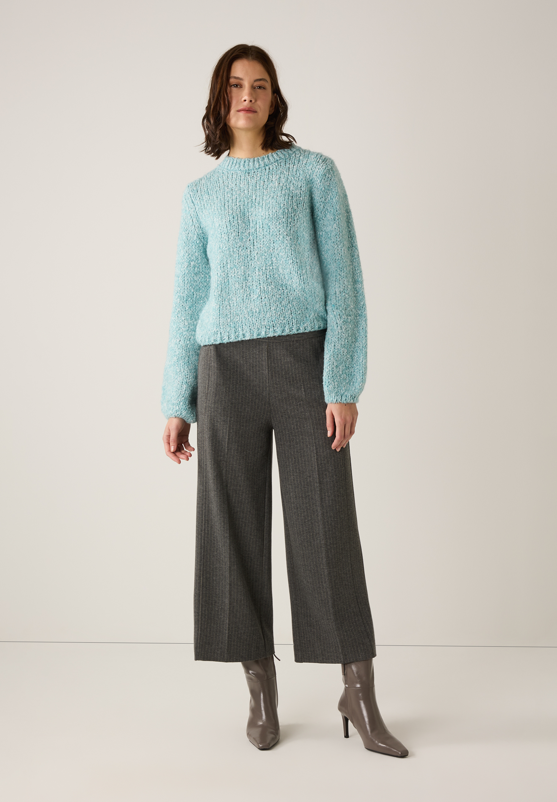 Eine Frau in MORE & MORE Strickware, einem blauen Bouclé-Pullover, grauer Hose und Stiefeln vor hellem Hintergrund. Eine Frau in MORE & MORE Strickware, einem blauen Bouclé-Pullover, grauer Hose und Stiefeln vor hellem Hintergrund.