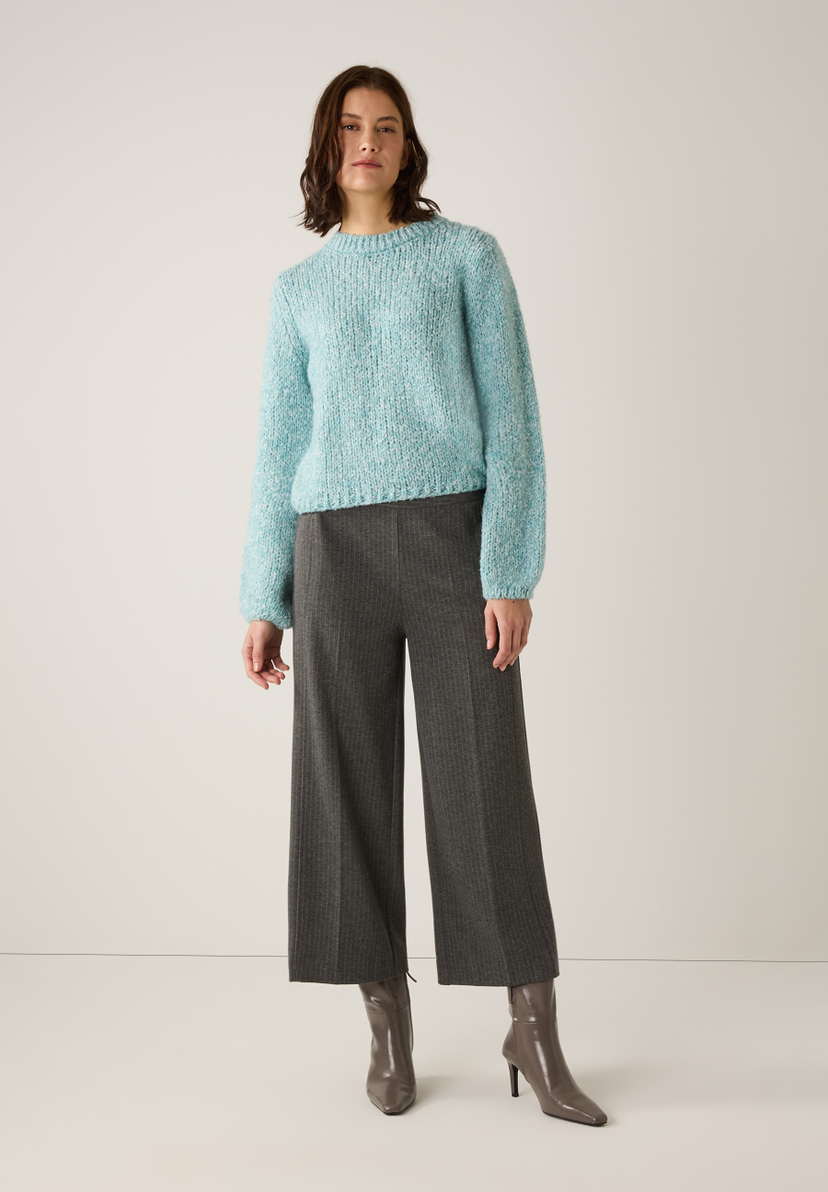 Eine Frau in MORE & MORE Strickware, einem blauen Bouclé-Pullover, grauer Hose und Stiefeln vor hellem Hintergrund. Eine Frau in MORE & MORE Strickware, einem blauen Bouclé-Pullover, grauer Hose und Stiefeln vor hellem Hintergrund.