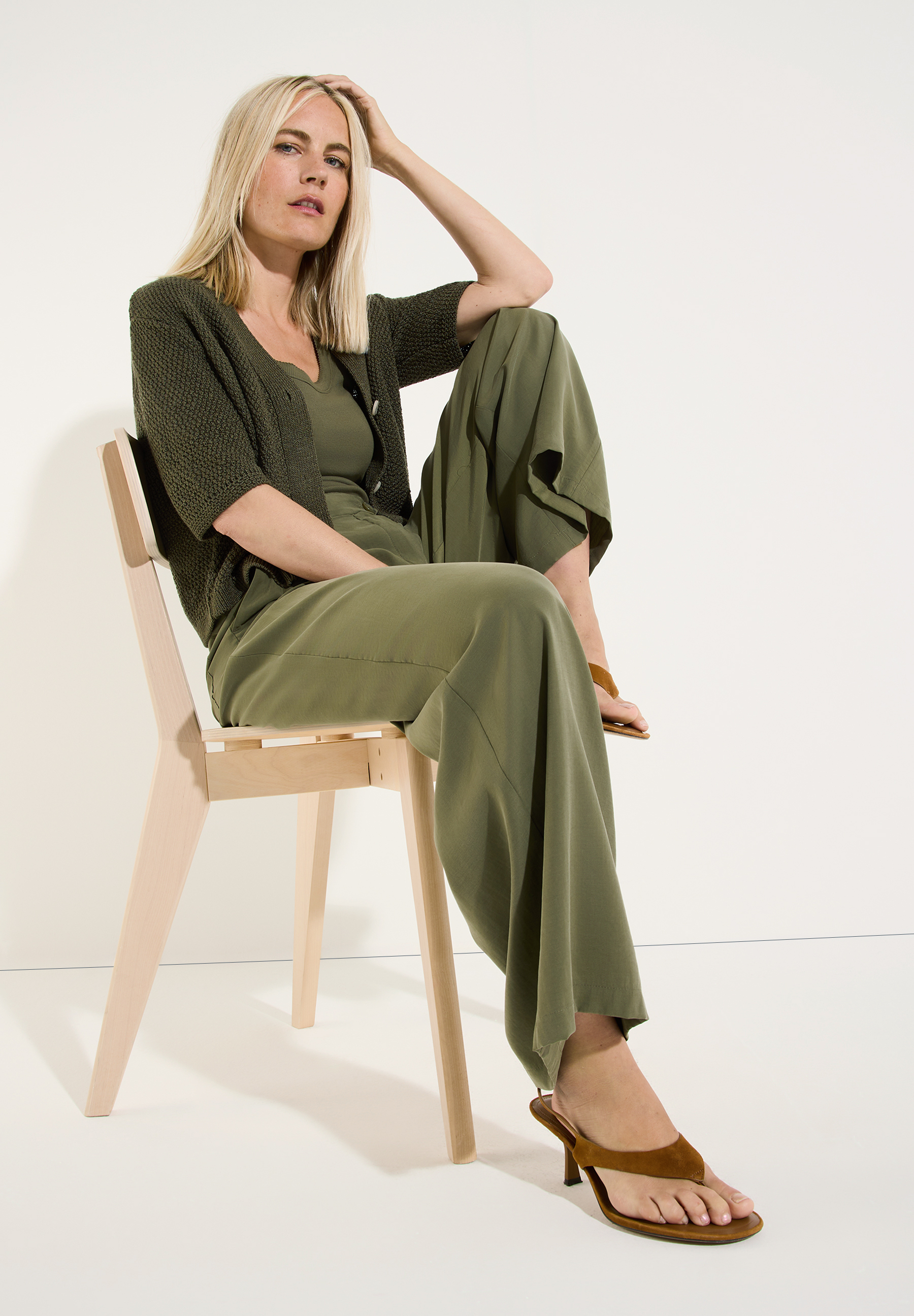 MORE & MORE Pullover & Strickmode: Blonde Frau in Khaki-Outfit sitzt seitlich auf hellem Holzstuhl. MORE & MORE Pullover & Strickmode: Blonde Frau in Khaki-Outfit sitzt seitlich auf hellem Holzstuhl.