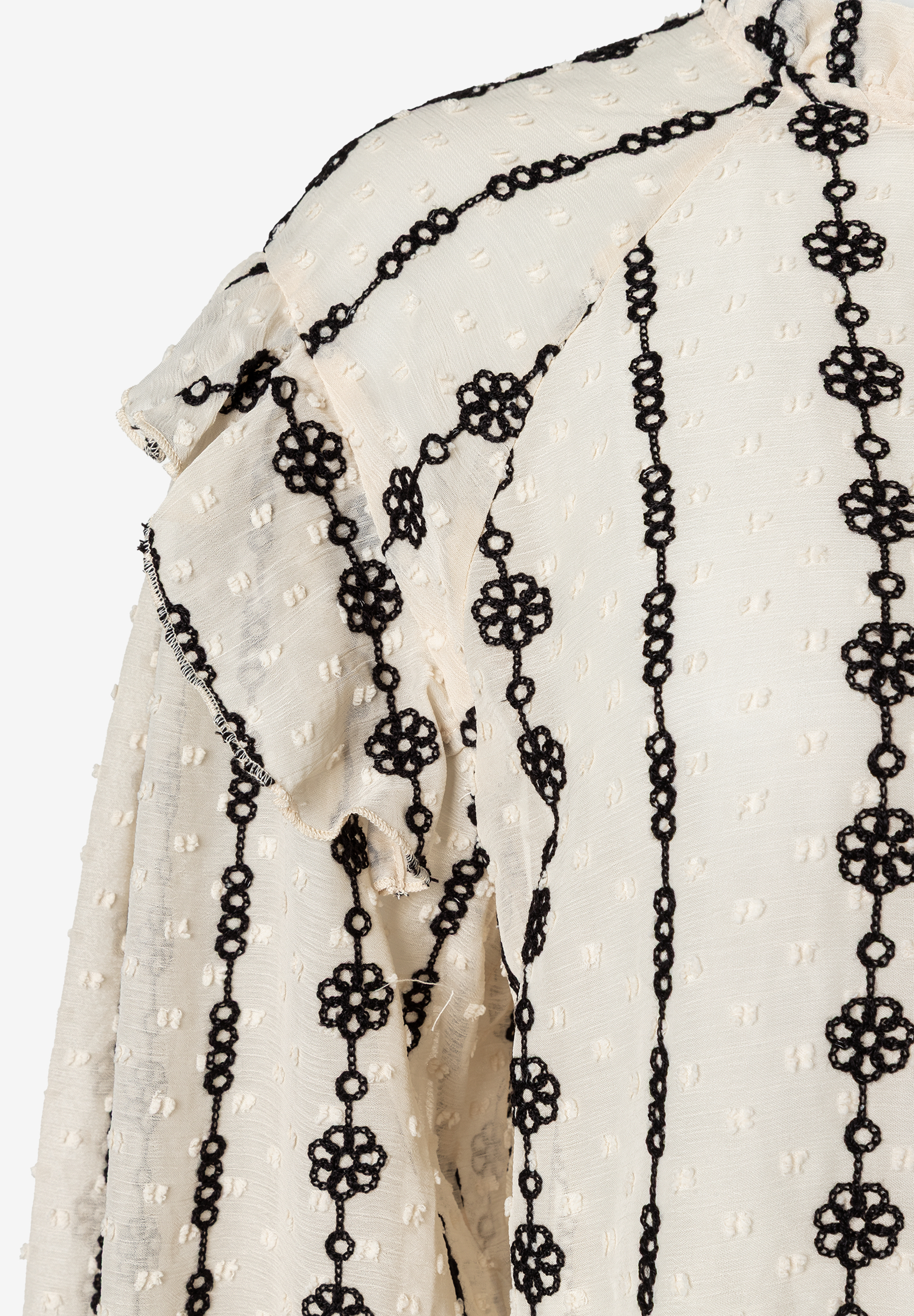 Nahaufnahme einer MORE & MORE Bluse: Cremefarbener Stoff mit schwarzen, blumenartigen Stickereien und Rüschen. Nahaufnahme einer MORE & MORE Bluse: Cremefarbener Stoff mit schwarzen, blumenartigen Stickereien und Rüschen.