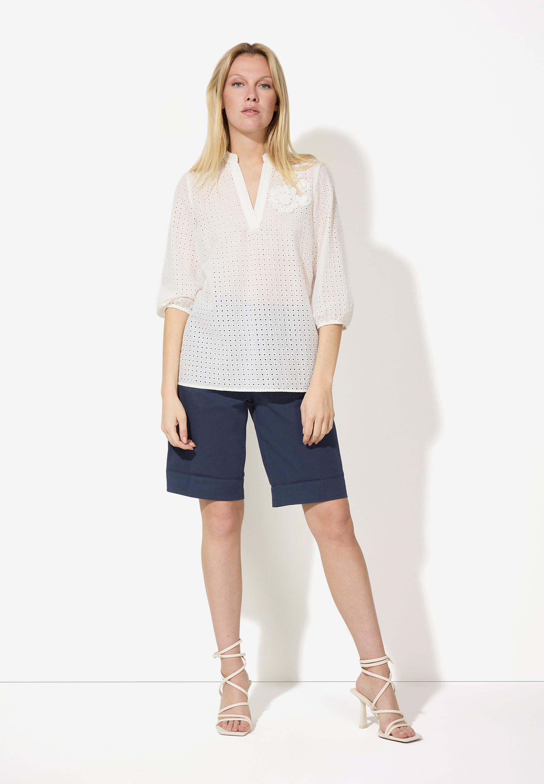 Eine Frau trägt eine weiße MORE & MORE Bluse mit Spitzenmaterial und blaue Shorts vor einem weißen Hintergrund. Eine Frau trägt eine weiße MORE & MORE Bluse mit Spitzenmaterial und blaue Shorts vor einem weißen Hintergrund.