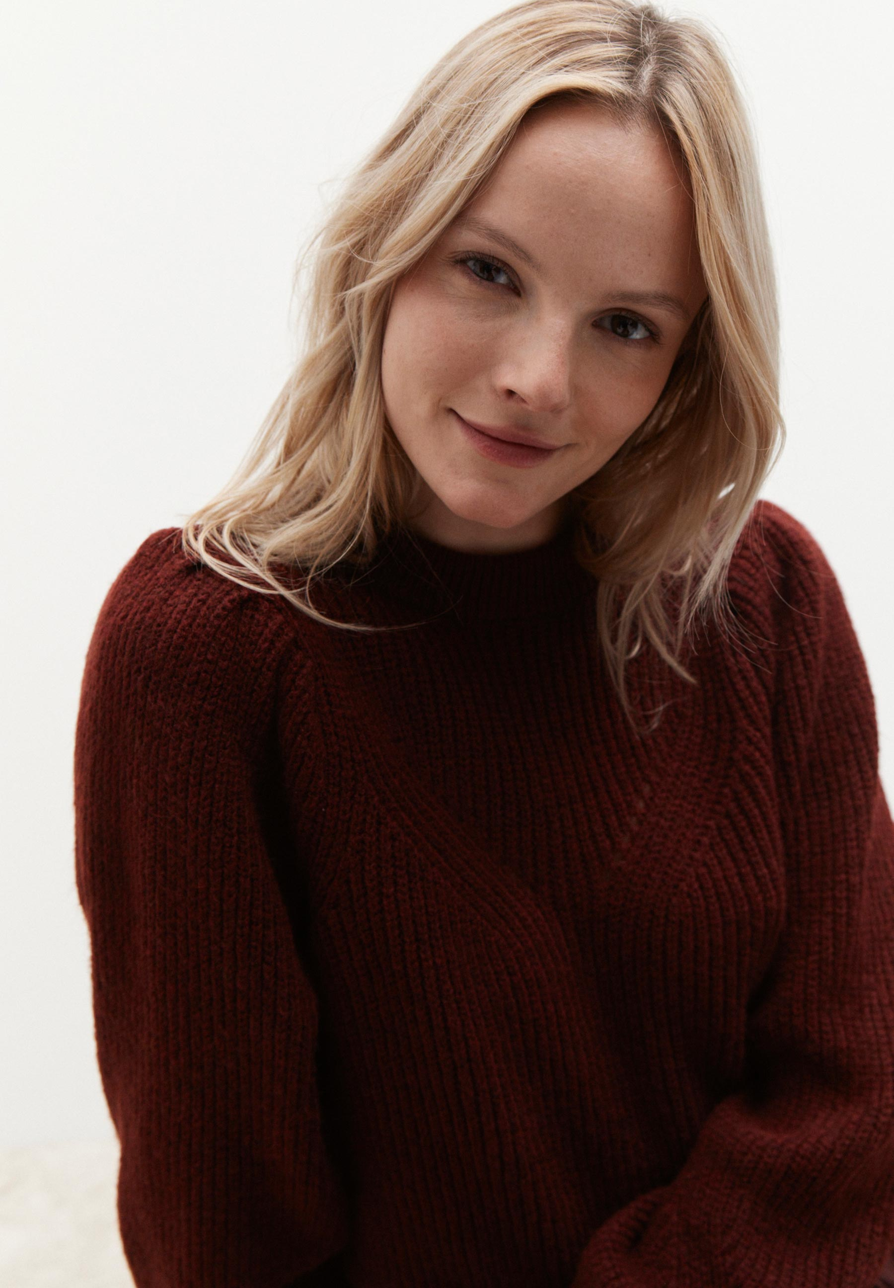 Eine Frau mit blonden, gewellten Haaren trägt einen bordeauxroten MORE & MORE Strickpullover und lächelt.