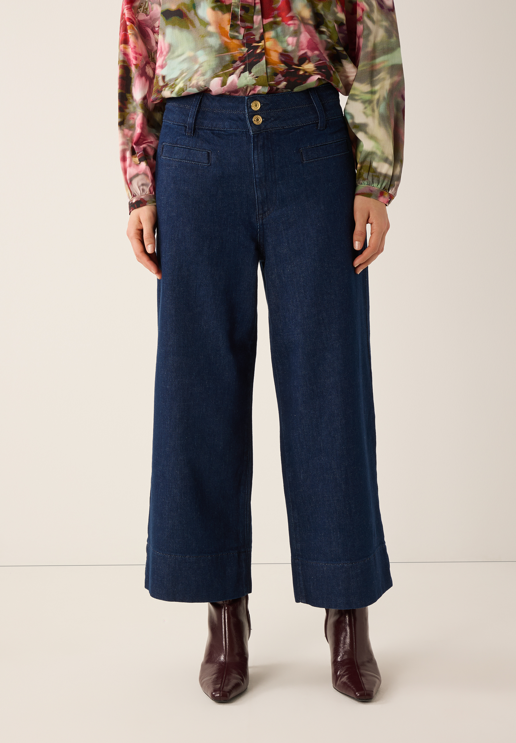 Eine Frau mit MORE & MORE Jeans und floraler Bluse steht vor einem neutralen Hintergrund.