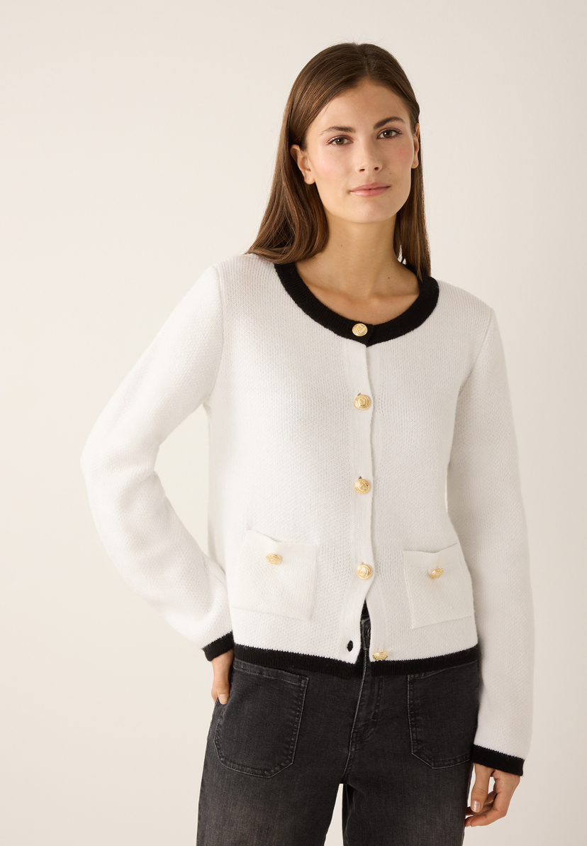 Eine Frau mit MORE & MORE Strickjacke. Die weiße Jacke hat goldene Knöpfe und schwarze Akzente an Kragen und Taschen. Eine Frau mit MORE & MORE Strickjacke. Die weiße Jacke hat goldene Knöpfe und schwarze Akzente an Kragen und Taschen.