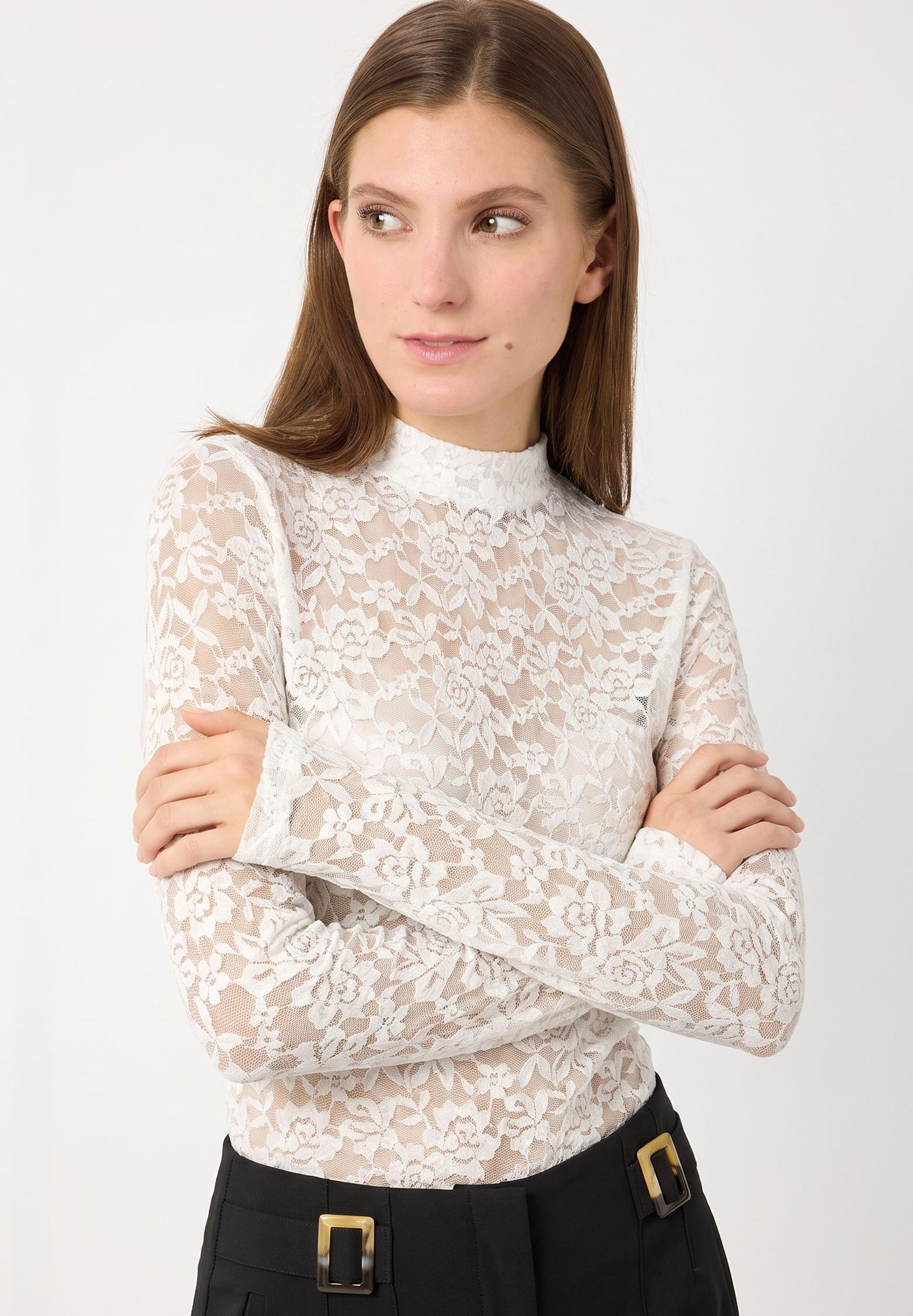 MORE & MORE Top aus elastischer weißer Spitze mit Blumenmuster, Rundhals und langen Ärmeln. Eine junge Frau in Schwarz trägt das Oberteil.