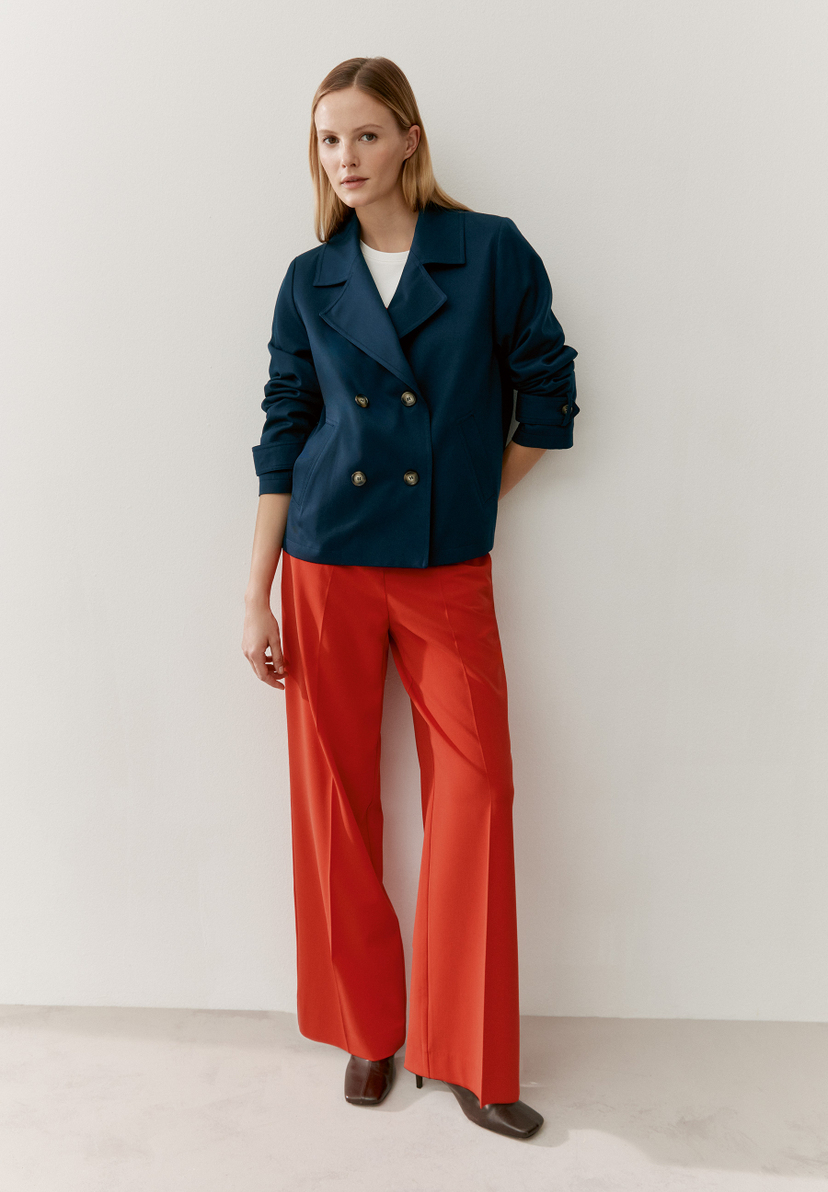 Eine Frau in einem MORE & MORE Oberteil und roter Hose posiert an einer weißen Wand. Eine Frau in einem MORE & MORE Oberteil und roter Hose posiert an einer weißen Wand.