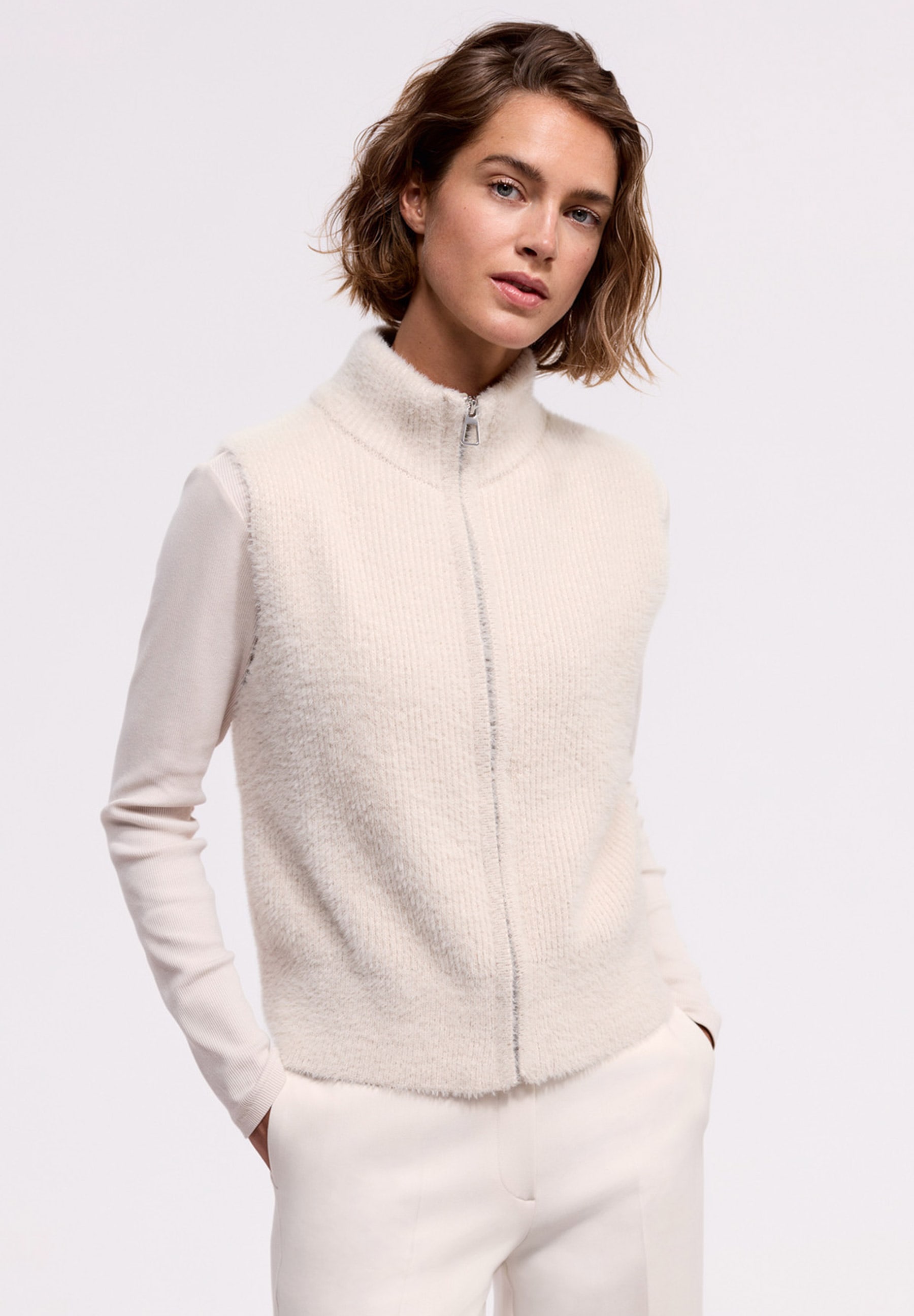 MORE & MORE Pullover: Eine Frau in einem creme-weißen Strickpullover mit Reißverschluss und passender Hose, Hände in den Taschen.