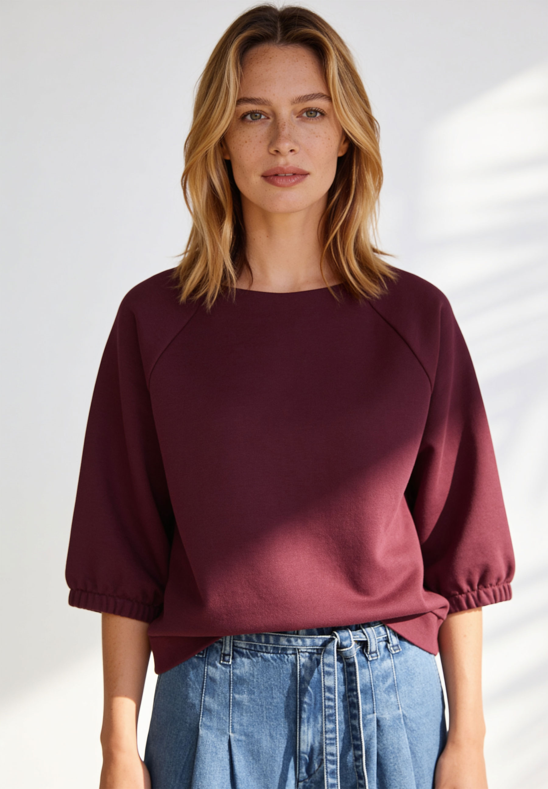 AI generated - MORE & MORE Damen-Sweatshirt in Beere. Das Model trägt ein weinrotes Oberteil mit 3/4-Ärmeln und Jeansrock.