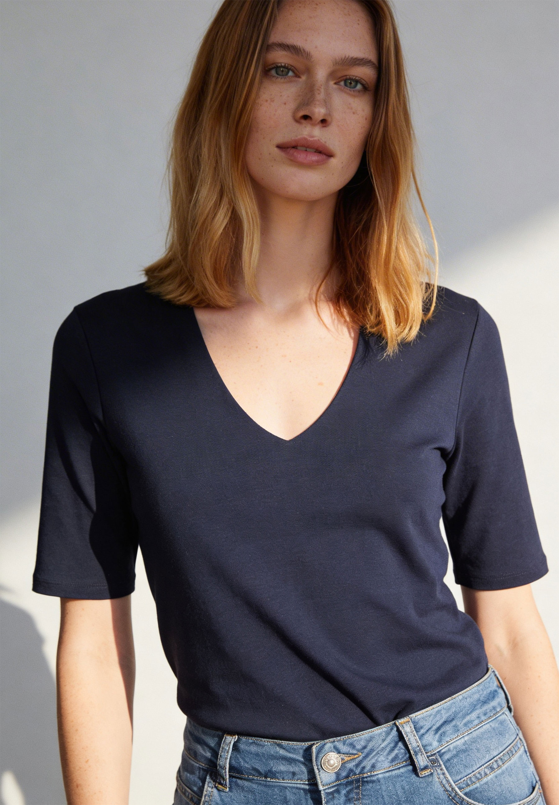 Eine Frau trägt ein marineblaues MORE & MORE Jersey-Top mit V-Ausschnitt und Jeans. Eine Frau trägt ein marineblaues MORE & MORE Jersey-Top mit V-Ausschnitt und Jeans.