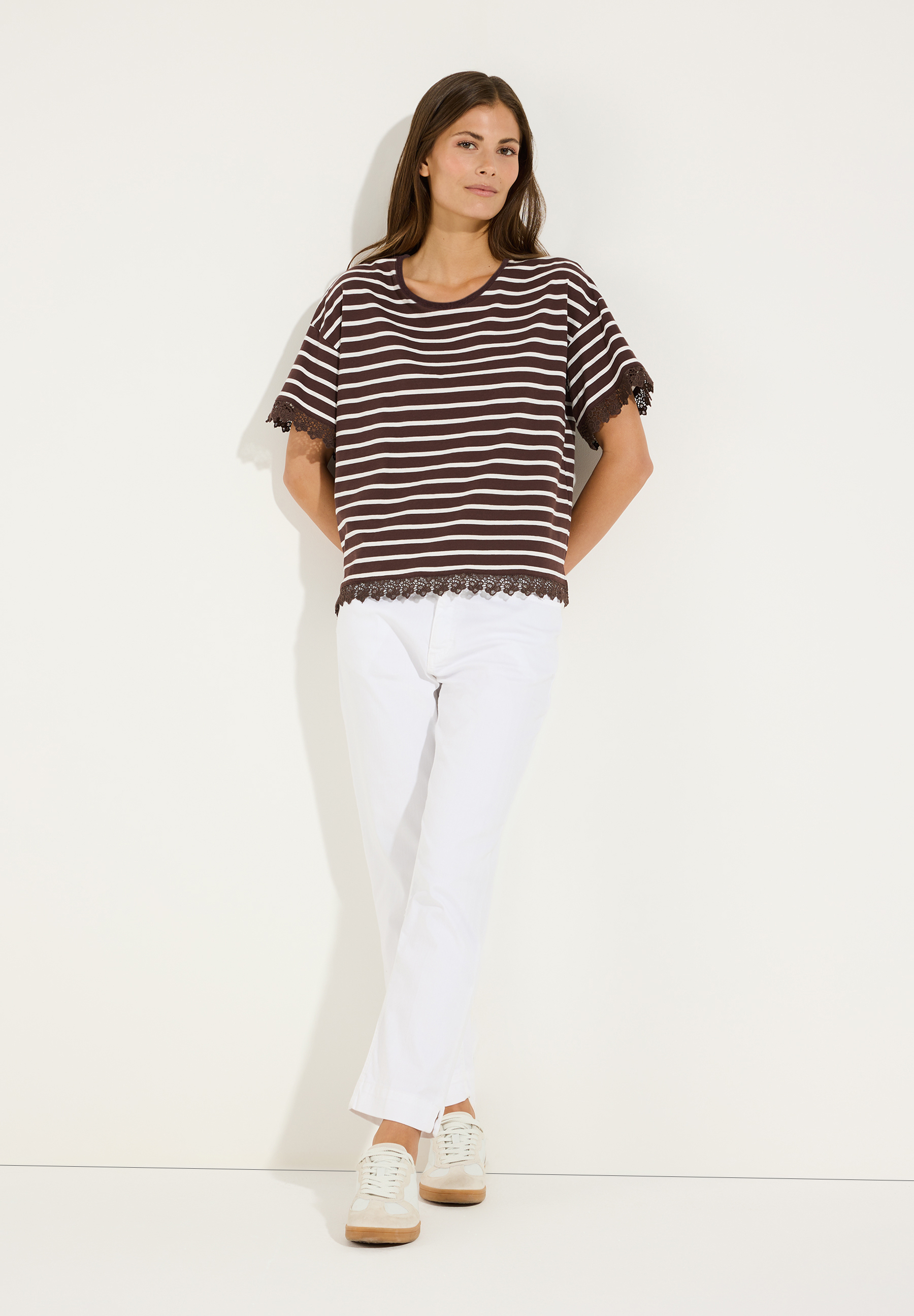 MORE & MORE Damen-Jersey-Shirt gestreift, mit Spitze an Ärmeln und Saum, in Braun/Weiß. Die Frau trägt dazu weiße Hosen und beigefarbene Sneaker. MORE & MORE Damen-Jersey-Shirt gestreift, mit Spitze an Ärmeln und Saum, in Braun/Weiß. Die Frau trägt dazu weiße Hosen und beigefarbene Sneaker.