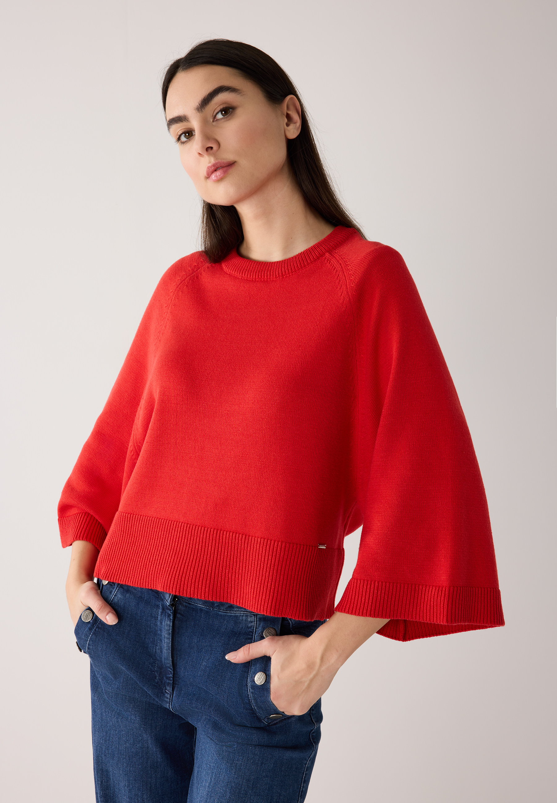 Eine Frau mit einem roten Strickpullover von MORE & MORE und Jeans mit Knöpfen an den Taschen.