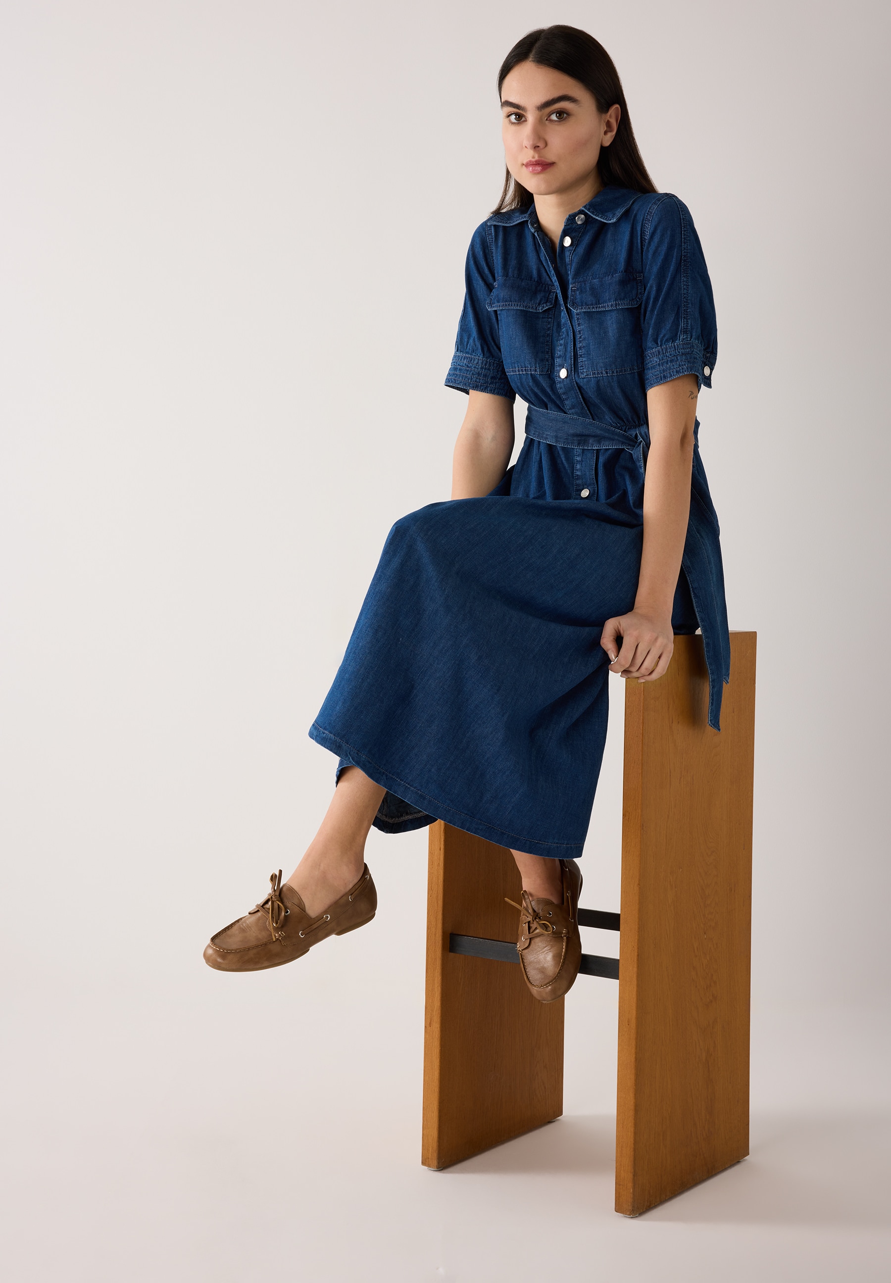 Eine Frau im MORE & MORE Jeanskleid sitzt auf einem Barhocker. Sie trägt braune Schuhe. Das Kleid hat einen Gürtel und kurze Ärmel. Eine Frau im MORE & MORE Jeanskleid sitzt auf einem Barhocker. Sie trägt braune Schuhe. Das Kleid hat einen Gürtel und kurze Ärmel.