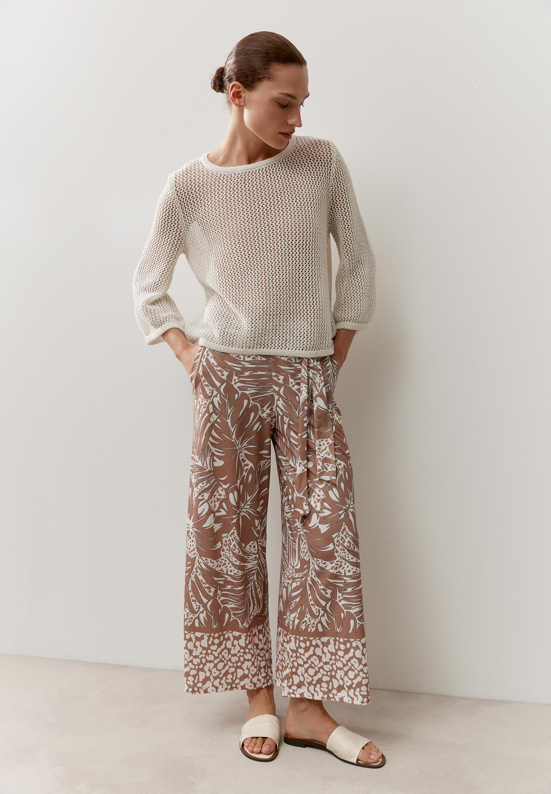 Eine Frau mit einem transparenten MORE & MORE Lochstrick-Pullover mit beigen Hosen mit Blattmuster. Eine Frau mit einem transparenten MORE & MORE Lochstrick-Pullover mit beigen Hosen mit Blattmuster.