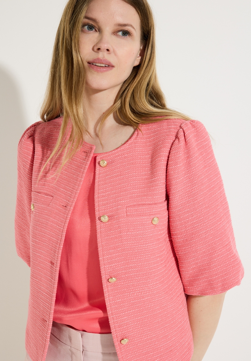 Eine Frau mit Bouclé-Blazer von MORE & MORE und beigen Hosen posiert vor einer weißen Wand. Eine Frau mit Bouclé-Blazer von MORE & MORE und beigen Hosen posiert vor einer weißen Wand.
