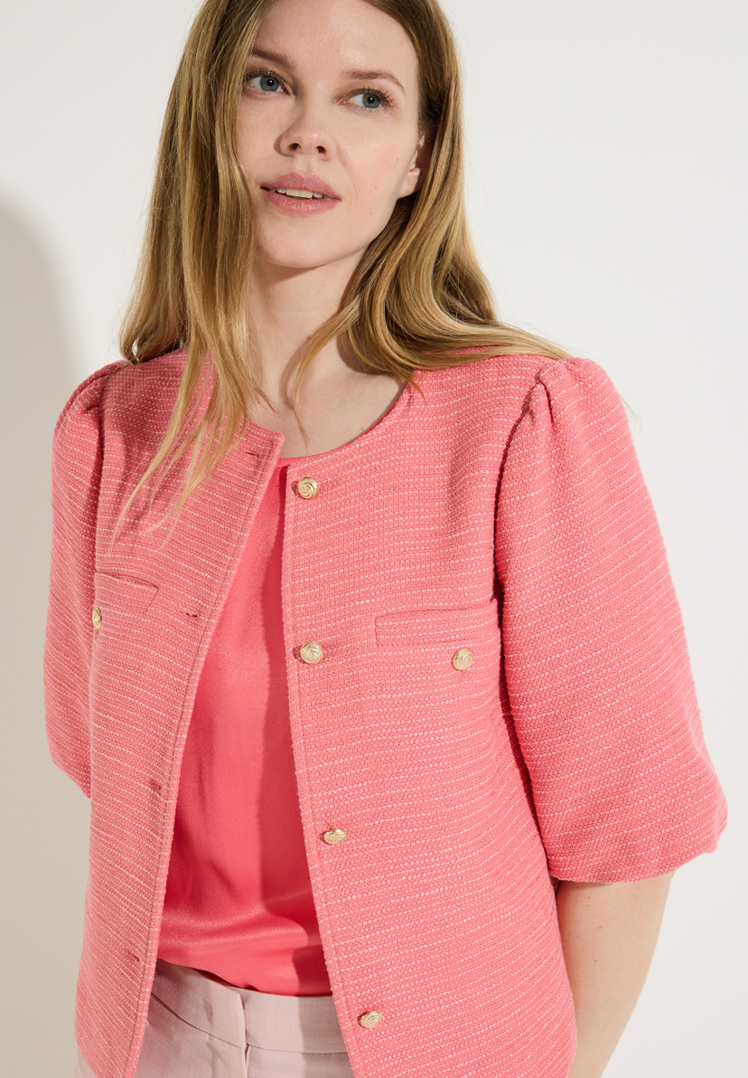 Eine Frau mit Bouclé-Blazer von MORE & MORE und beigen Hosen posiert vor einer weißen Wand. Eine Frau mit Bouclé-Blazer von MORE & MORE und beigen Hosen posiert vor einer weißen Wand.