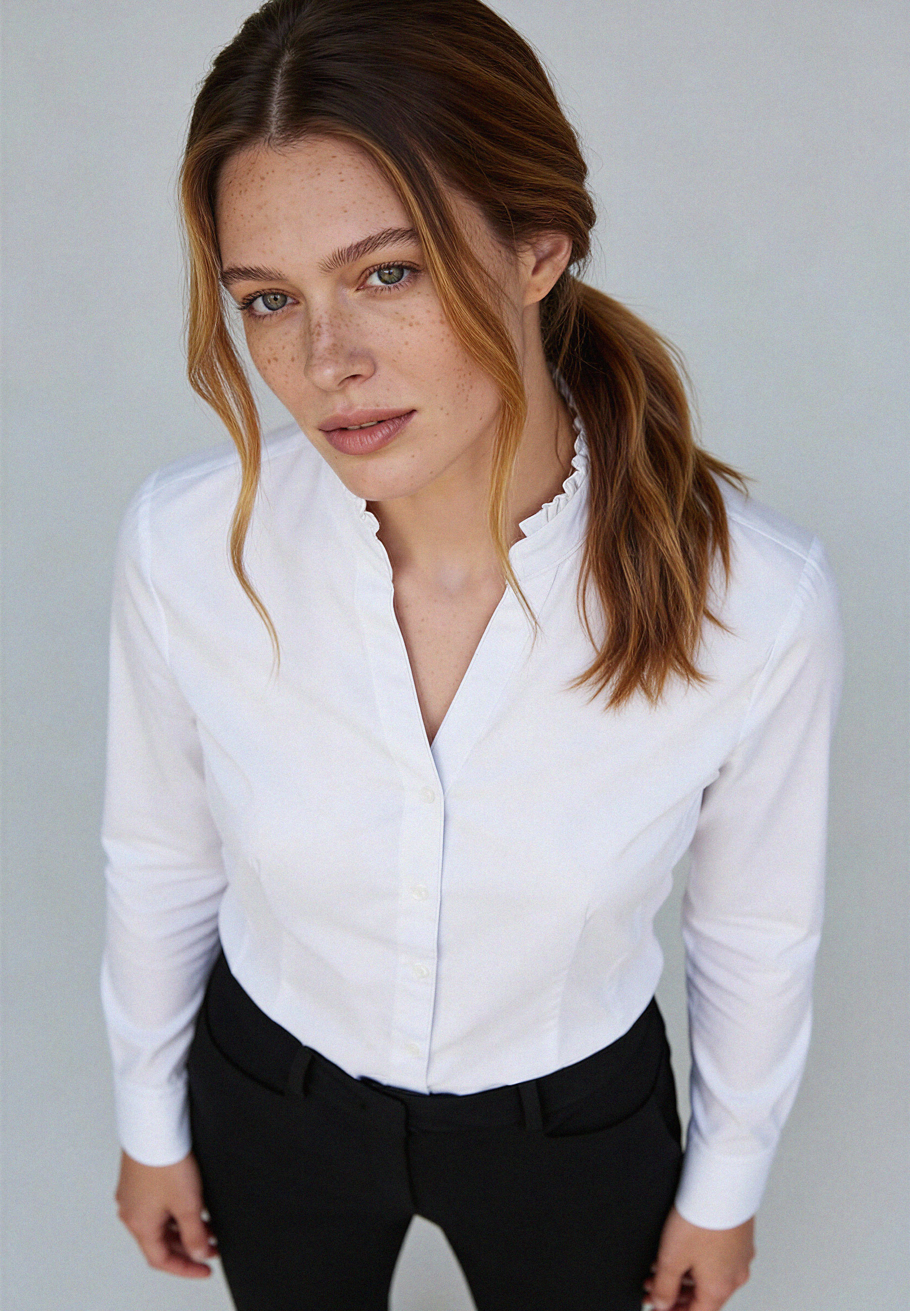 Eine Frau mit Sommersprossen trägt eine weiße MORE & MORE Bluse aus Baumwollmischung und eine schwarze Hose. Blick von oben. Eine Frau mit Sommersprossen trägt eine weiße MORE & MORE Bluse aus Baumwollmischung und eine schwarze Hose. Blick von oben.