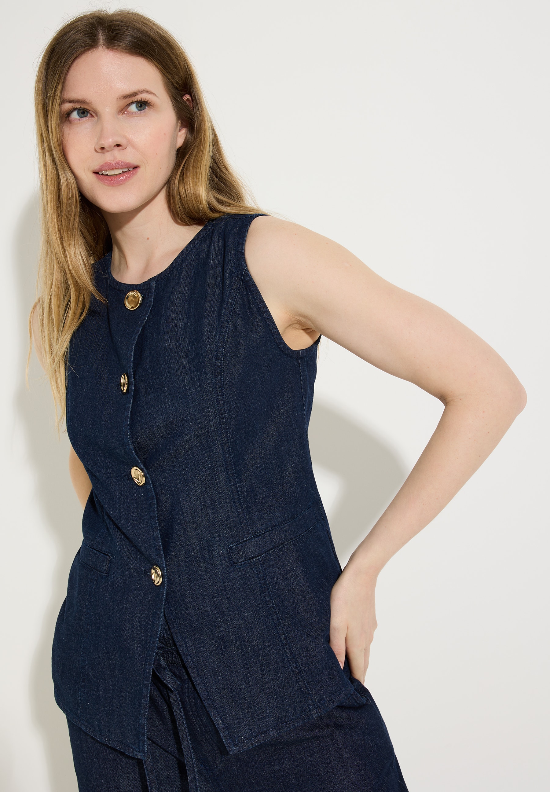 Eine Frau trägt ein ärmelloses MORE & MORE Oberteil mit goldenen Knöpfen.