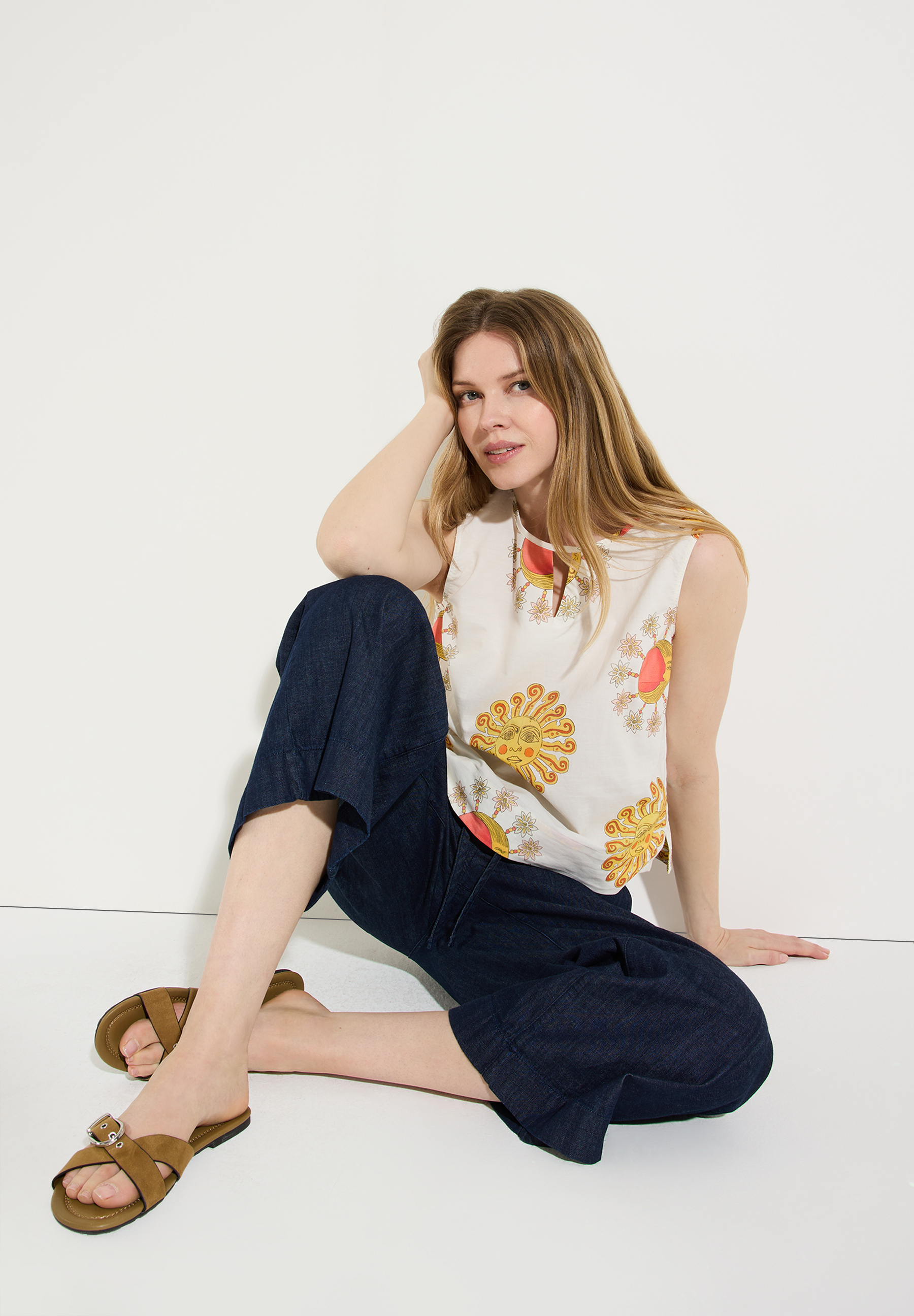 Eine Frau sitzt mit einer Bluse von MORE & MORE mit Sonnen-Motiven und dunklen Jeans auf dem Boden. Eine Frau sitzt mit einer Bluse von MORE & MORE mit Sonnen-Motiven und dunklen Jeans auf dem Boden.